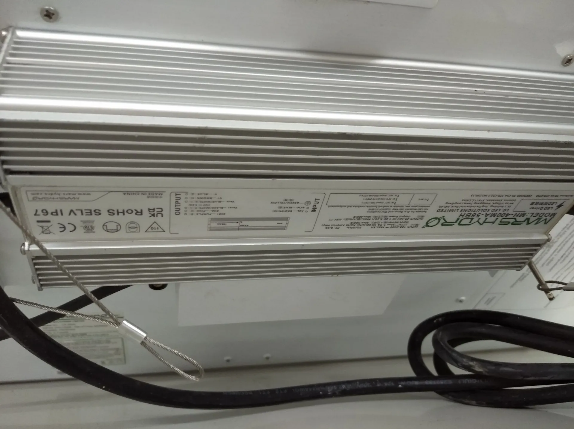 Srebrny, aluminiowy zasilacz LED z radiatorowymi żebrami. Na obudowie znajduje się biała tabliczka znamionowa z tekstem, w tym napisy: 'UK RoHS SELV IP67', 'MARSHYRO', 'MODEL: MH-400MA-56BH'. Do urządzenia podłączone są czarne kable elektryczne oraz cienka stalowa linka zabezpieczająca.