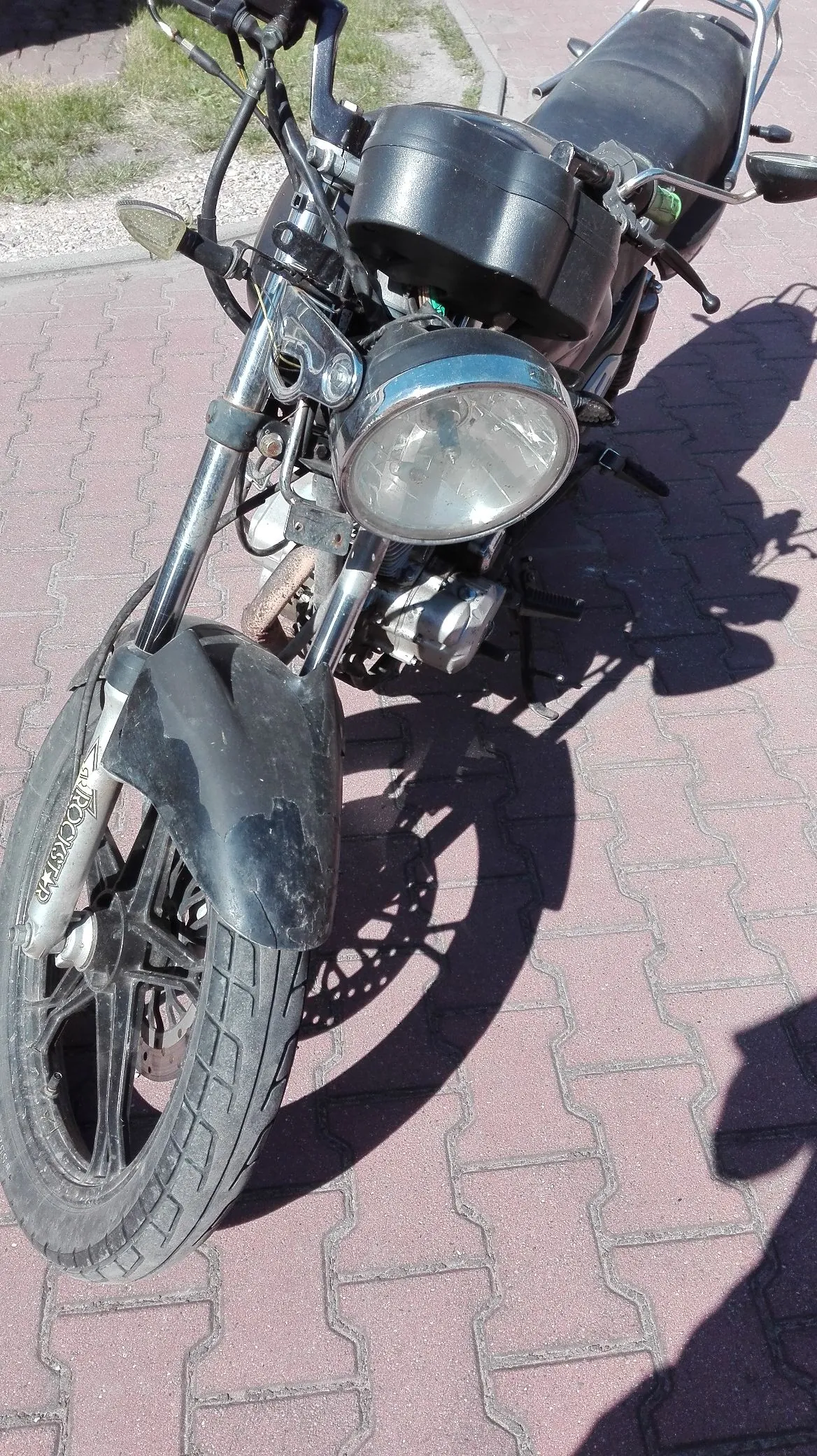 Zdjęcie motocykla zaparkowanego na czerwonym chodniku z kostki brukowej. Widoczny jest przód pojazdu z dużym, okrągłym reflektorem, przednim błotnikiem z napisem 'ROCKSTER' oraz przednim kołem. Motocykl jest czarny, a słońce rzuca wyraźny cień na nawierzchnię.