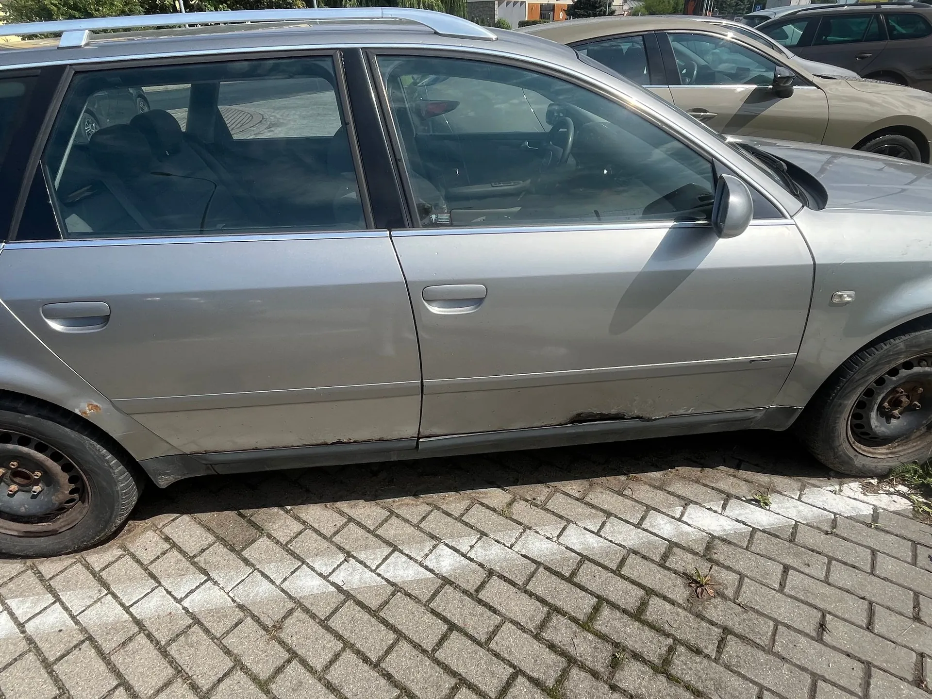Boczny widok srebrnego samochodu typu kombi zaparkowanego na kostce brukowej. Na dolnej krawędzi drzwi i progu widoczne są ślady korozji i rdzy. W tle widać fragmenty innych samochodów.