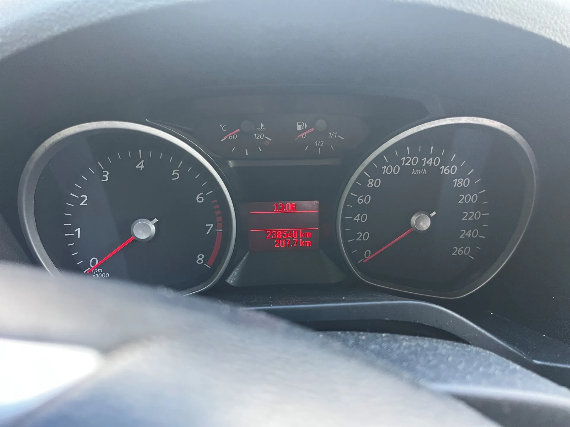 Panel instrumentów w samochodzie. Po lewej stronie znajduje się obrotomierz z czerwoną wskazówką, po prawej prędkościomierz z podziałką do 260 km/h. W centralnej części znajduje się cyfrowy wyświetlacz z czerwonym tekstem: godzina '13:08', przebieg '238540 km' oraz dystans '207.7 km'. Nad wyświetlaczem widoczne są kontrolki temperatury silnika i poziomu paliwa.