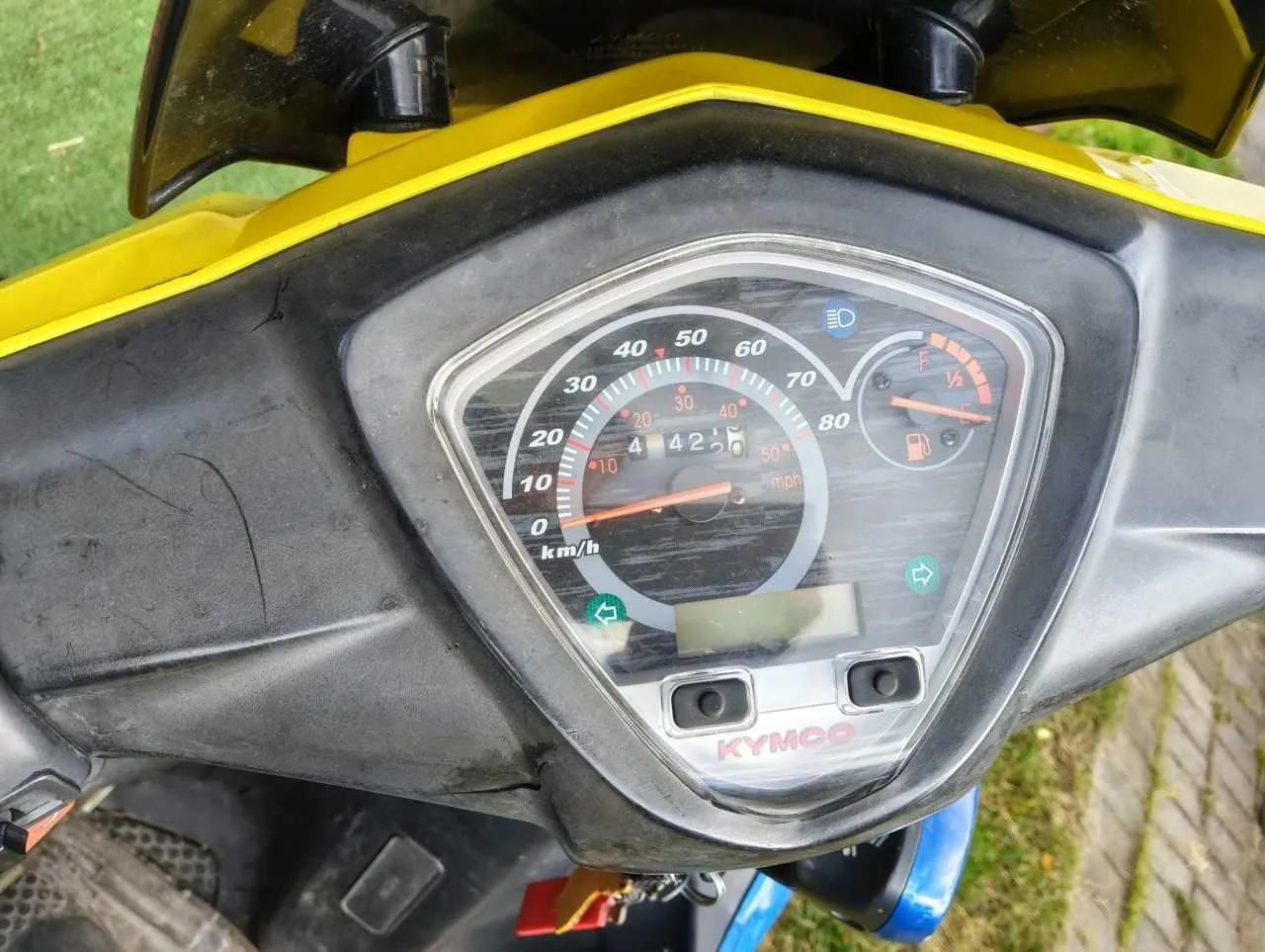 Zbliżenie na deskę rozdzielczą żółtego skutera marki KYMCO. Centralnym elementem jest analogowy prędkościomierz z podziałką w km/h (od 0 do 80) oraz mniejszą podziałką w mph. Obok znajduje się wskaźnik poziomu paliwa oraz wskaźnik temperatury. Na dole obudowy widnieje napis KYMCO.