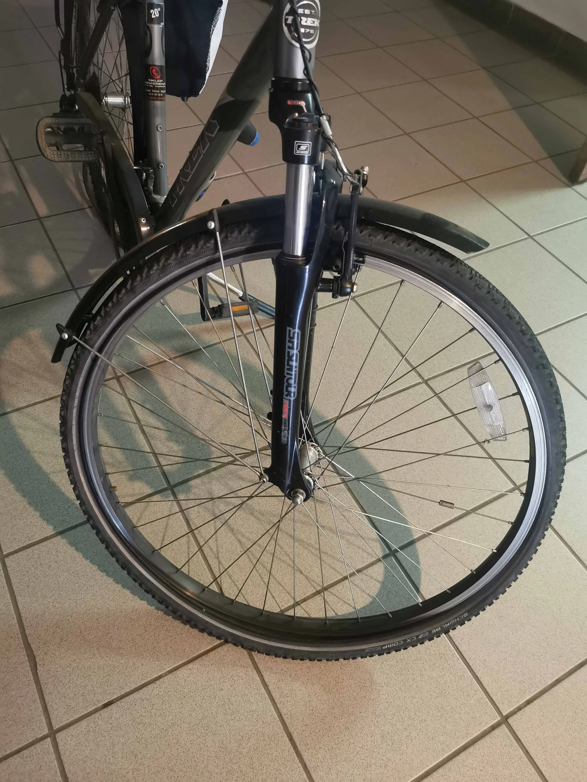 Zbliżenie na przednie koło i widelec amortyzowany roweru trekkingowego. Rower stoi na jasnej, kafelkowej podłodze. Na widelcu widnieje napis 'SR SUNTOUR'. Koło posiada czarną oponę z bieżnikiem, srebrne szprychy oraz zamontowany nad kołem czarny błotnik.