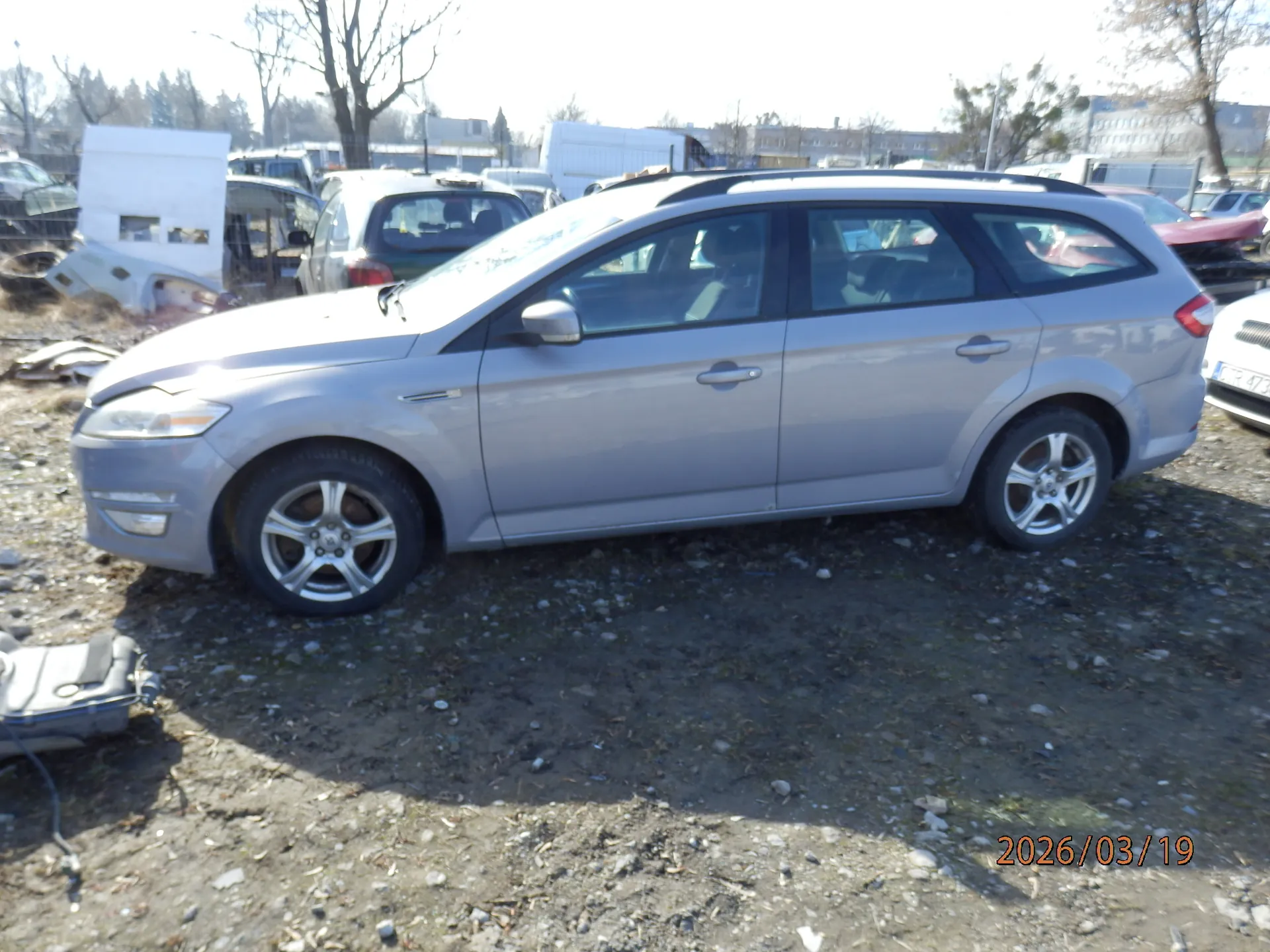 Srebrny samochód typu kombi zaparkowany na nierównym, żwirowym terenie. W tle widoczne są inne pojazdy, w tym częściowo zdemontowane lub uszkodzone, białe budowle, drzewa bez liści oraz budynki. W prawym dolnym rogu obrazu znajduje się pomarańczowy napis z datą 2026/03/19. Słońce oświetla scenę, rzucając cienie na ziemię.