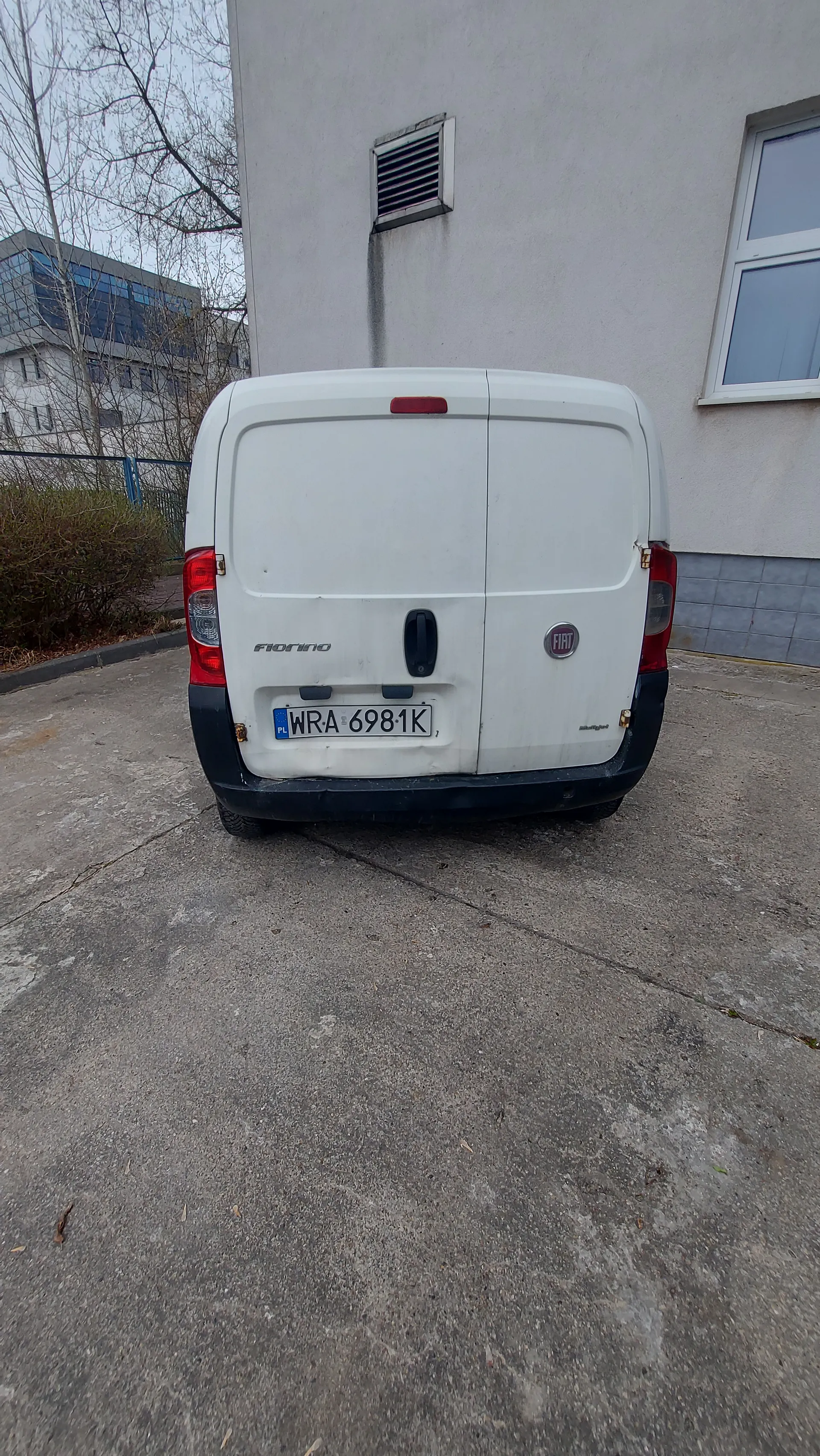 Biały samochód dostawczy Fiat Fiorino zaparkowany tyłem do obserwatora. Widoczny jest biały tył pojazdu z czerwonymi światłami, logo Fiata, napisem "Fiorino" oraz tablicą rejestracyjną WRA 6981K z niebieskim pasem PL. Po lewej stronie obrazu budynek z oknem i wentylacją, na dole betonowa powierzchnia, w tle drzewa bez liści i inny budynek. Tekst na tablicy rejestracyjnej jest pionowy, co wskazuje na nieprawidłową orientację obrazu.