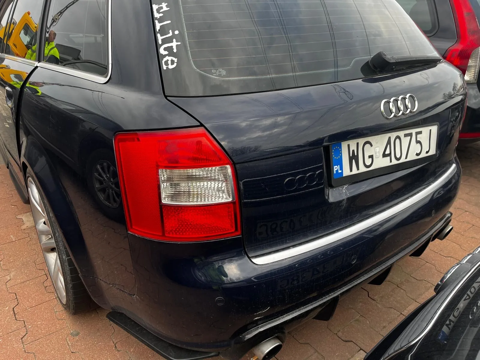 Tył czarnego samochodu marki Audi z tablicą rejestracyjną PL WG 4075J. Na tylnej szybie widoczny pionowy napis 'elite'. Samochód stoi na brukowanej powierzchni, obok częściowo widoczny inny pojazd. W tle osoba w żółtym kamizelce odblaskowej oraz fragment żółtego pojazdu. Czerwone światła tylnego reflektora i emblemat Audi są wyraźnie widoczne.