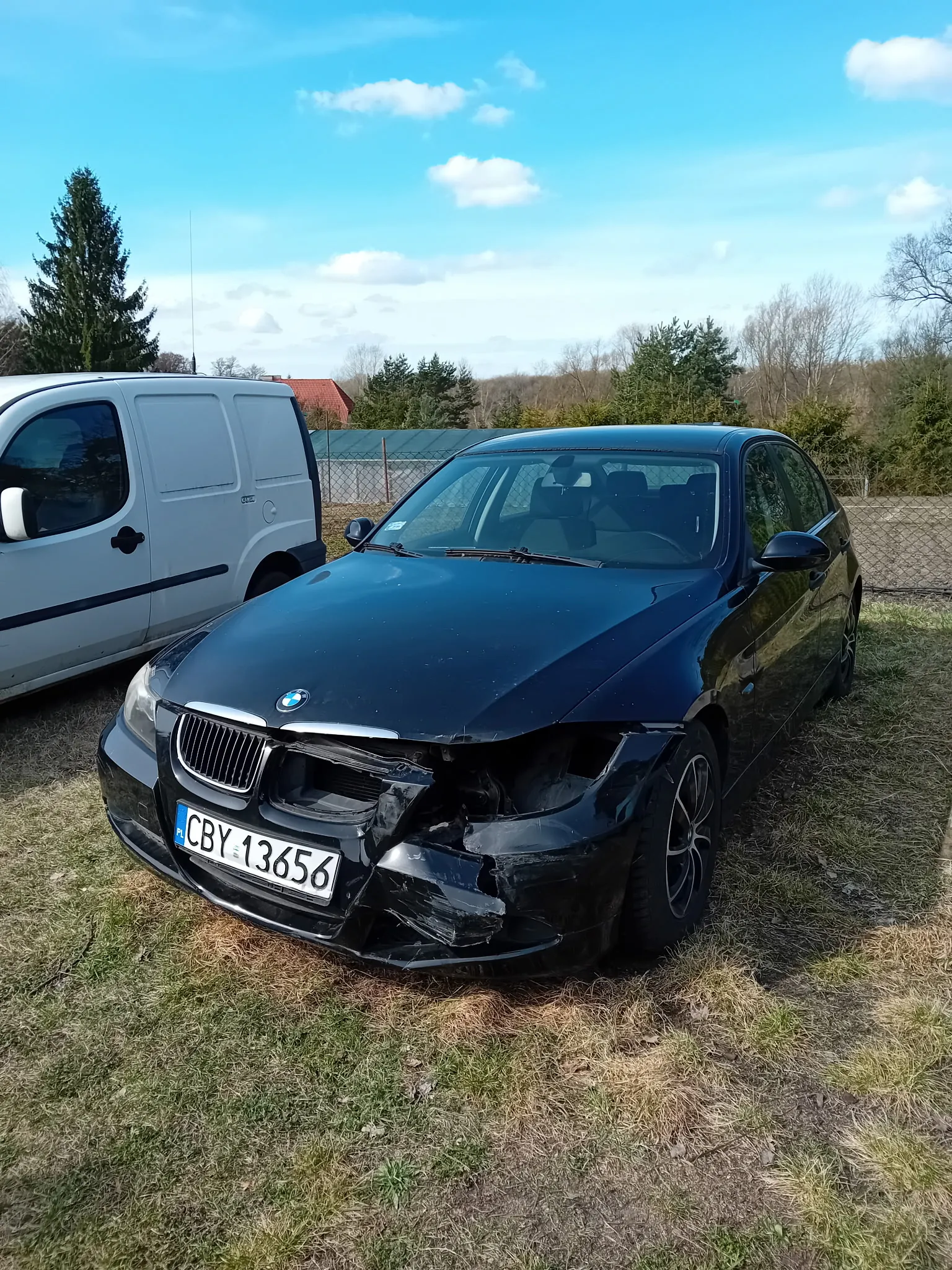 Czarny samochód marki BMW z uszkodzonym przodem, tablica rejestracyjna CBY 13656. Po lewej stronie biały furgon. Tło: trawa, drzewa, ogrodzenie, dom z czerwonym dachem i niebo z chmurami.