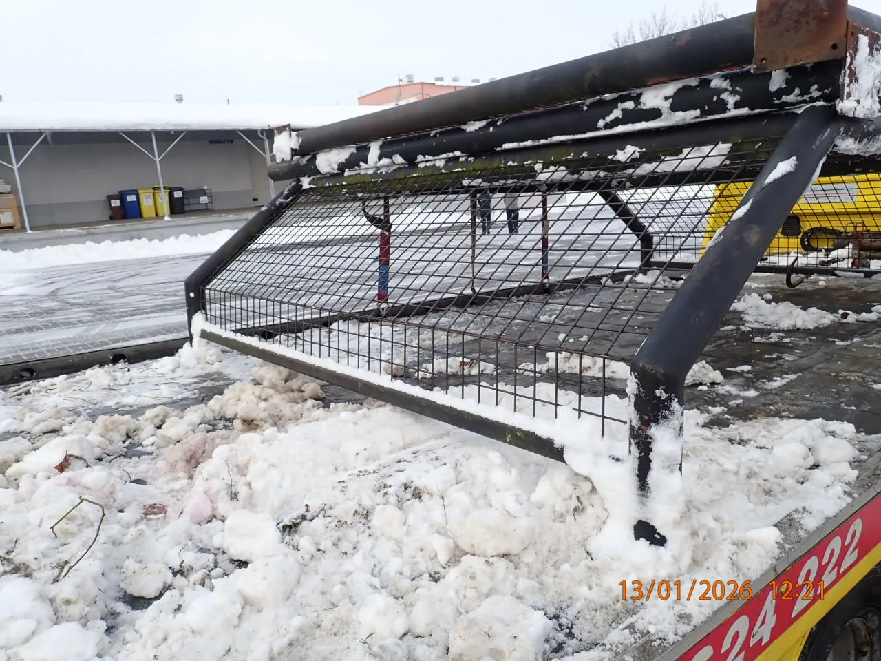 Zdjęcie przedstawia metalową siatkę z czarnymi rurami, pokrytą śniegiem, umocowaną na żółtym pojeździe z czerwonymi cyframi 24 222. W tle widoczny jest budynek z daszkiem, kolorowe pojemniki na śmieci (niebieski, żółty, czarny) oraz śnieżna powierzchnia. W prawym dolnym rogu znajduje się pomarańczowy znaczek z datą 13/01/2026 i godziną 12:21. Niebo jest pochmurne, a na ziemi leży rozproszony śnieg.