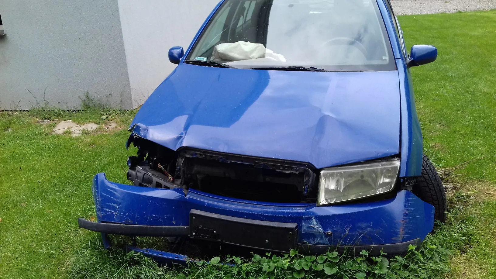 Niebieski samochód osobowy z poważnie uszkodzonym przodem. Maska jest wgnieciona, przedni zderzak jest częściowo oderwany i opada na trawę, a grill oraz lewa lampa są zniszczone lub brakujące. Wewnątrz pojazdu, za przednią szybą, widoczna jest rozwinięta biała poduszka powietrzna. Samochód stoi na zielonej trawie obok jasnej ściany budynku.