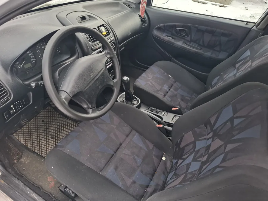 Wnętrze samochodu z widoczną kierownicą, deską rozdzielczą, dźwignią zmiany biegów i siedzeniami. Siedzenia mają czarny wzór z niebieskimi i szarymi trójkątami. Deska rozdzielcza zawiera wskaźniki prędkości i obrotomierza. Widoczne są również drzwi z uchwytem i okno, za którym znajduje się śnieg. Podłoga pokryta jest czarną siatkową wykładziną.