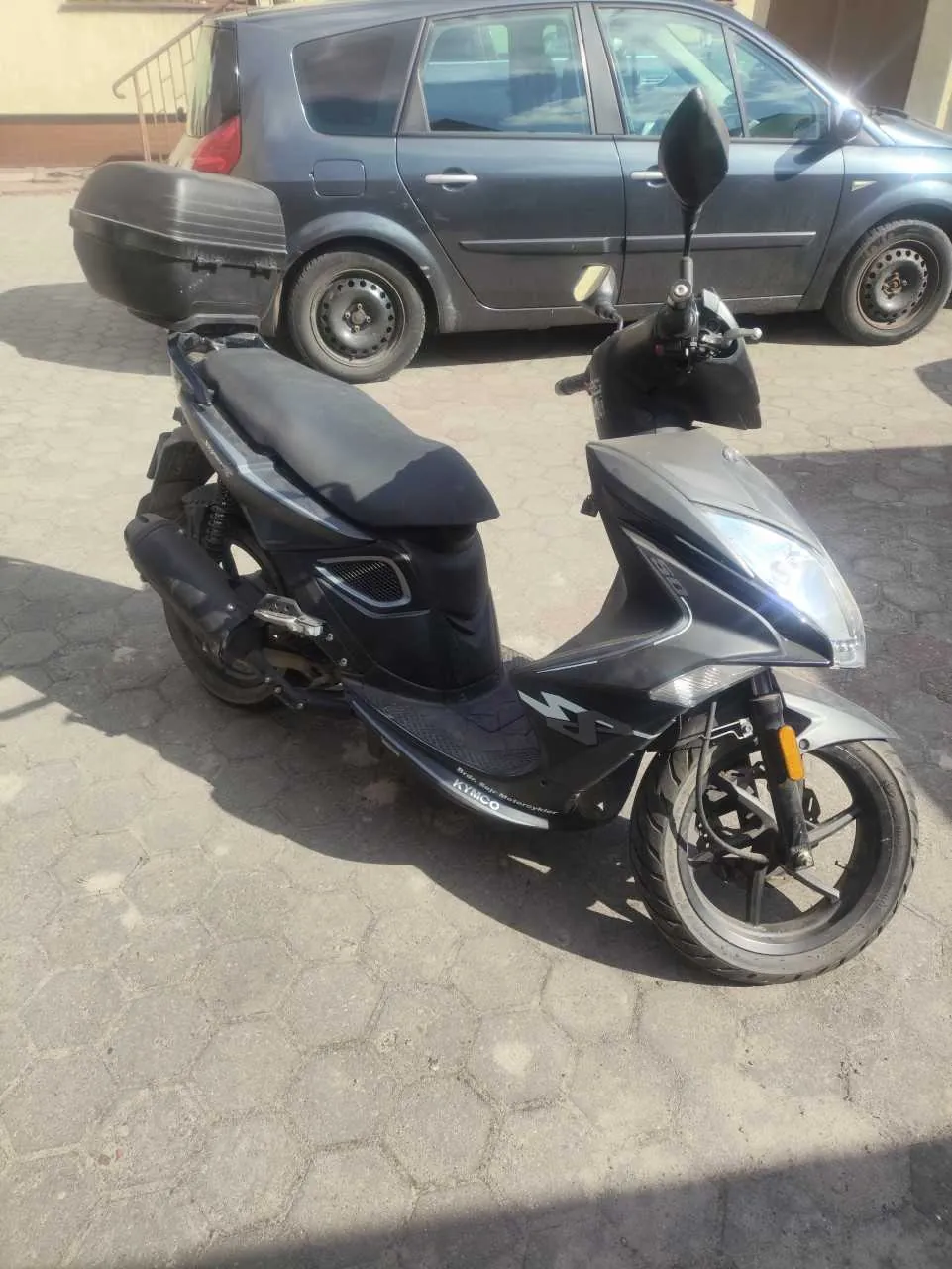 Czarno-srebrny skuter marki KYMCO zaparkowany na chodniku z sześciokątnymi płytami. W tle ciemnoszary samochód osobowy typu hatchback oraz fragment budynku z metalową balustradą. Słoneczny dzień, widoczne cienie na powierzchni chodnika.