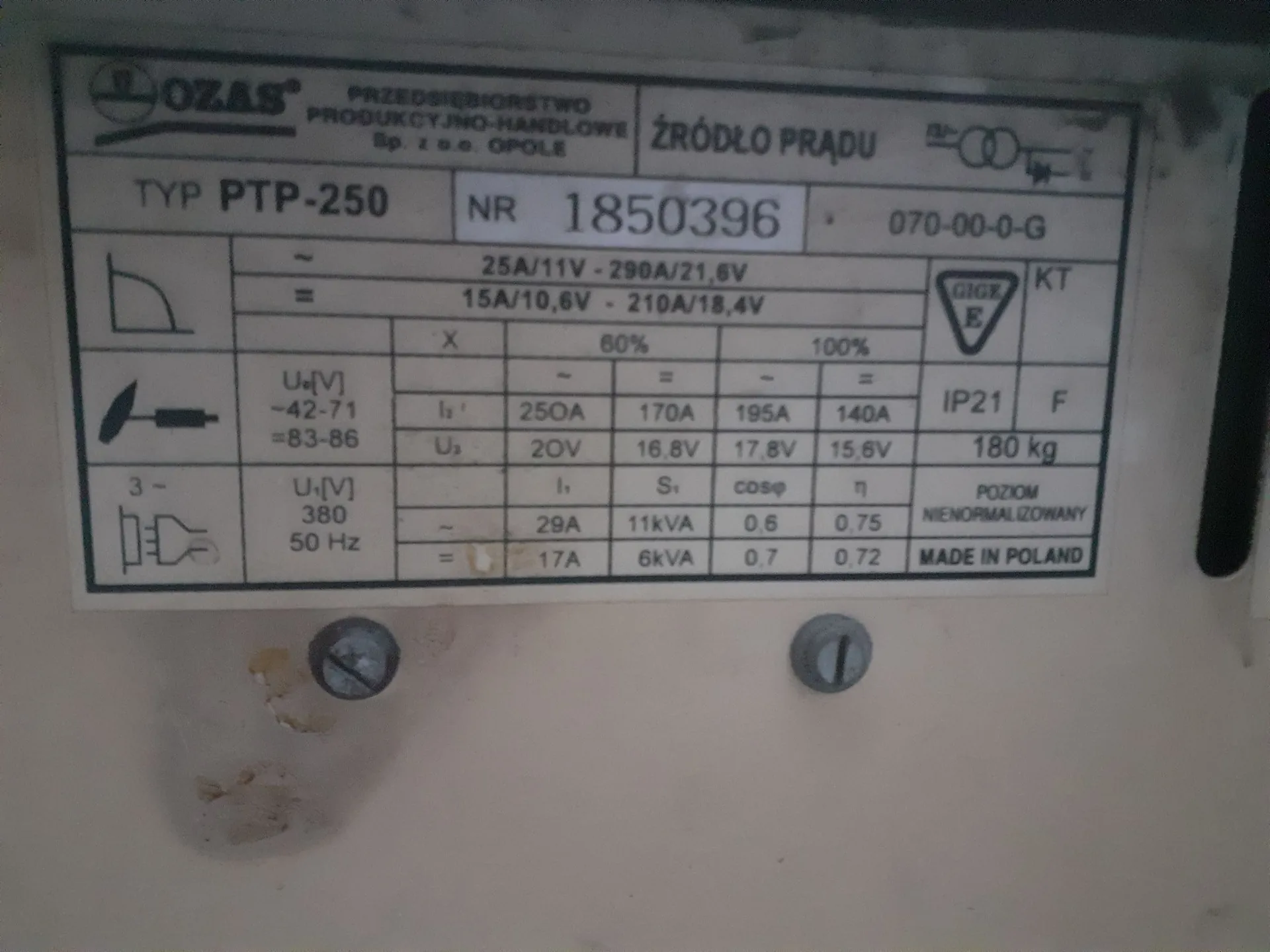 Tabliczka znamionowa urządzenia elektrycznego marki OZAS. Na górze widnieje napis: 'OZAS PRZEDSIĘBIORSTWO PRODUKCYJNO-HANDLOWE Sp. z o.o. OPOLE ŹRÓDŁO PRĄDU'. Poniżej znajdują się dane techniczne: TYP PTP-250, NR 1850396, kod 070-00-0-G. Tabela zawiera parametry prądowe i napięciowe, m.in. 25A/11V - 290A/21,6V oraz 15A/10,6V - 210A/18,4V. Widoczne są również oznaczenia IP21, masa 180 kg oraz napis 'MADE IN POLAND'. Tabliczka jest przykręcona do jasnej obudowy za pomocą dwóch śrub.