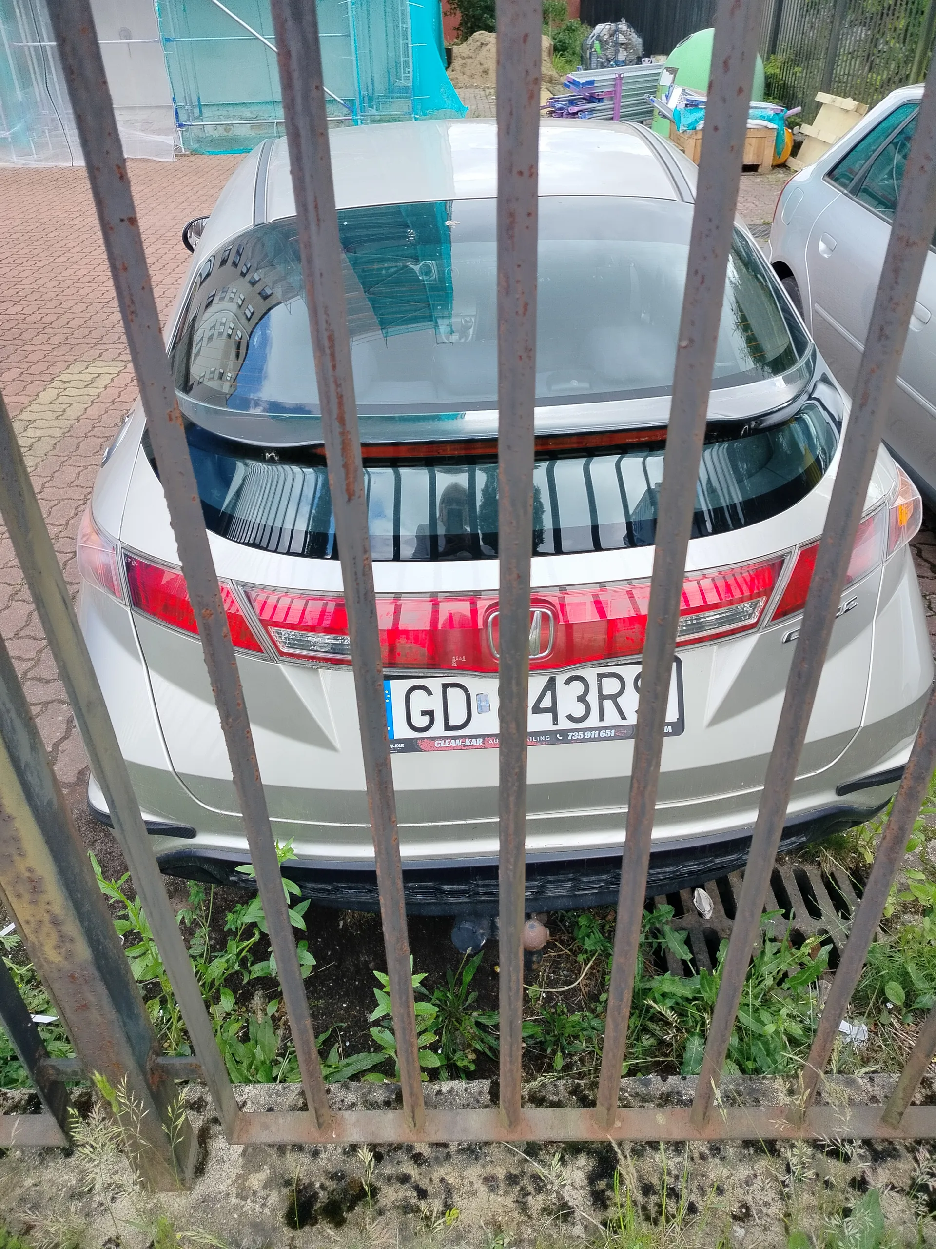 Zdjęcie przedstawia tył srebrnego samochodu marki Honda za rdzewiałym metalowym ogrodzeniem. Na tablicy rejestracyjnej widoczny jest numer GD 43RS oraz naklejka z napisem CLEAN-KAR. W tle znajduje się drugi srebrny samochód, budowa z niebieskim rusztowaniem oraz materiały budowlane. Pod ogrodzeniem rośnie trawa i widoczna jest studzienka kanalizacyjna.