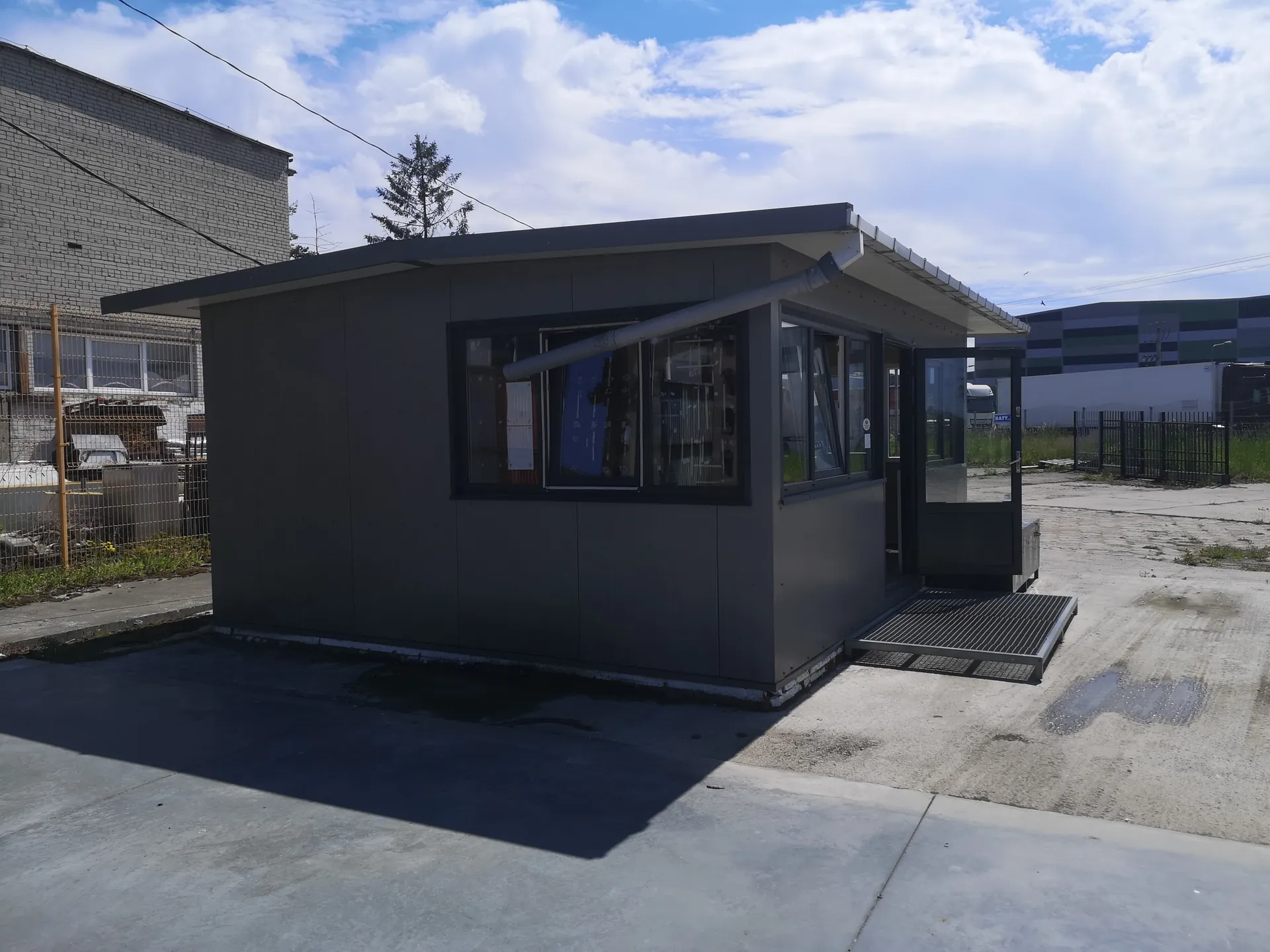 Mały szary budynek strażniczy lub kiosk z dużymi oknami i otwartymi drzwiami szklanymi. Budynek posiada dach z daszkiem, metalową rynną oraz rampę prowadzącą do wejścia. Znajduje się na betonowej powierzchni, w tle widoczne są ceglany budynek, przemysłowa hala z kolorowymi pasami, drzewo, ogrodzenie i częściowo zachmurzone niebo. Wokół rosną trawy i widoczne są plamy wilgoci na ziemi.