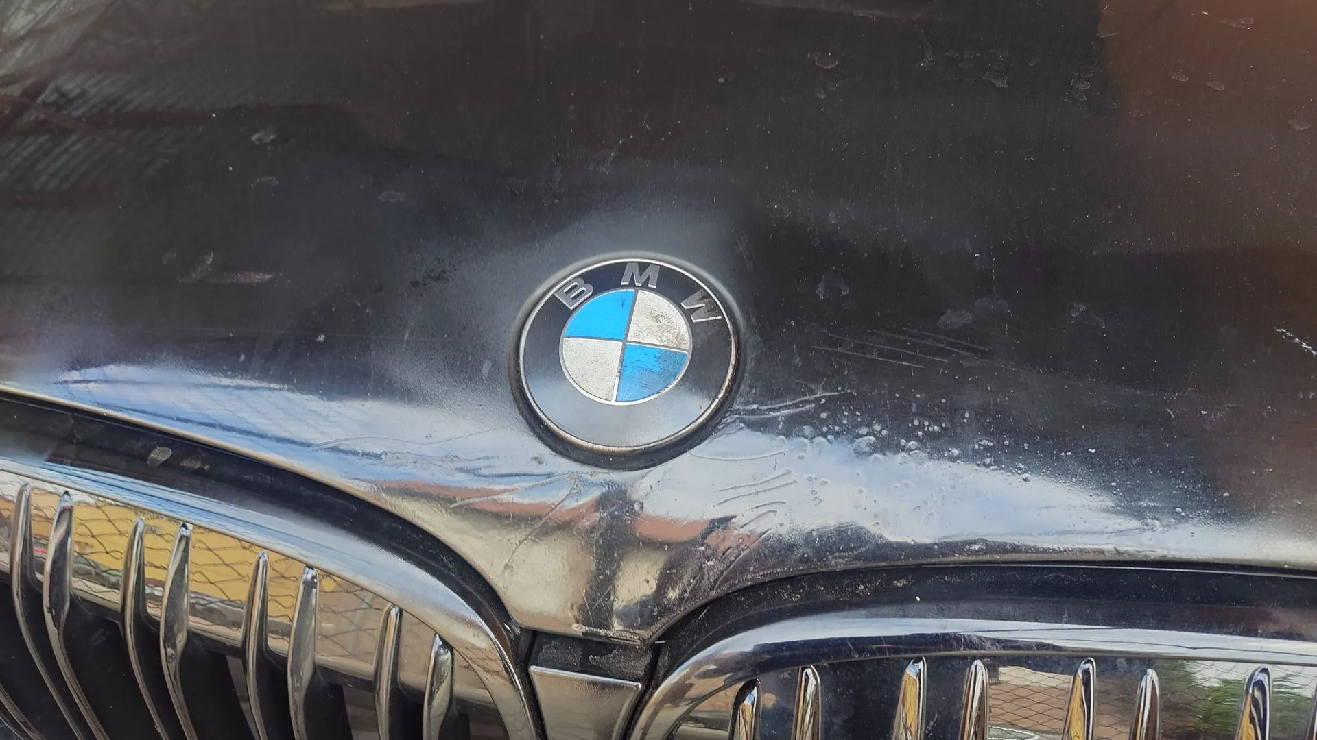 Zbliżenie na czarną maskę samochodu z centralnie umieszczonym logo marki BMW. Logo składa się z okręgu z napisem BMW na górnym, czarnym obrzeżu oraz niebiesko-białych pól wewnątrz. Powierzchnia lakieru jest zarysowana i zabrudzona. W dolnej części zdjęcia widoczny jest fragment czarnego grilla samochodu.