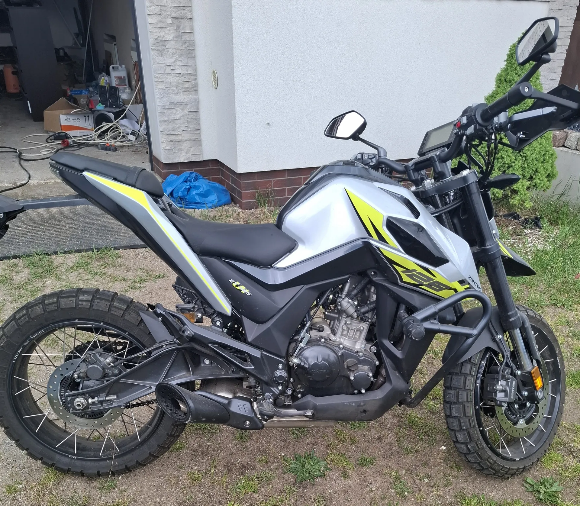 Motocykl Zontes 125 w kolorze srebrnym, czarnym i żółtym, zaparkowany na podwórku z żwirem i trawą. W tle budynek z kamiennej i białej elewacji, otwarty garaż z widocznymi pudełkami, przewodami i niebieską folią. Widoczne elementy motocykla: czarne siedzenie, lusterka, tablica rejestracyjna, silnik i opony z głębokim profilem.