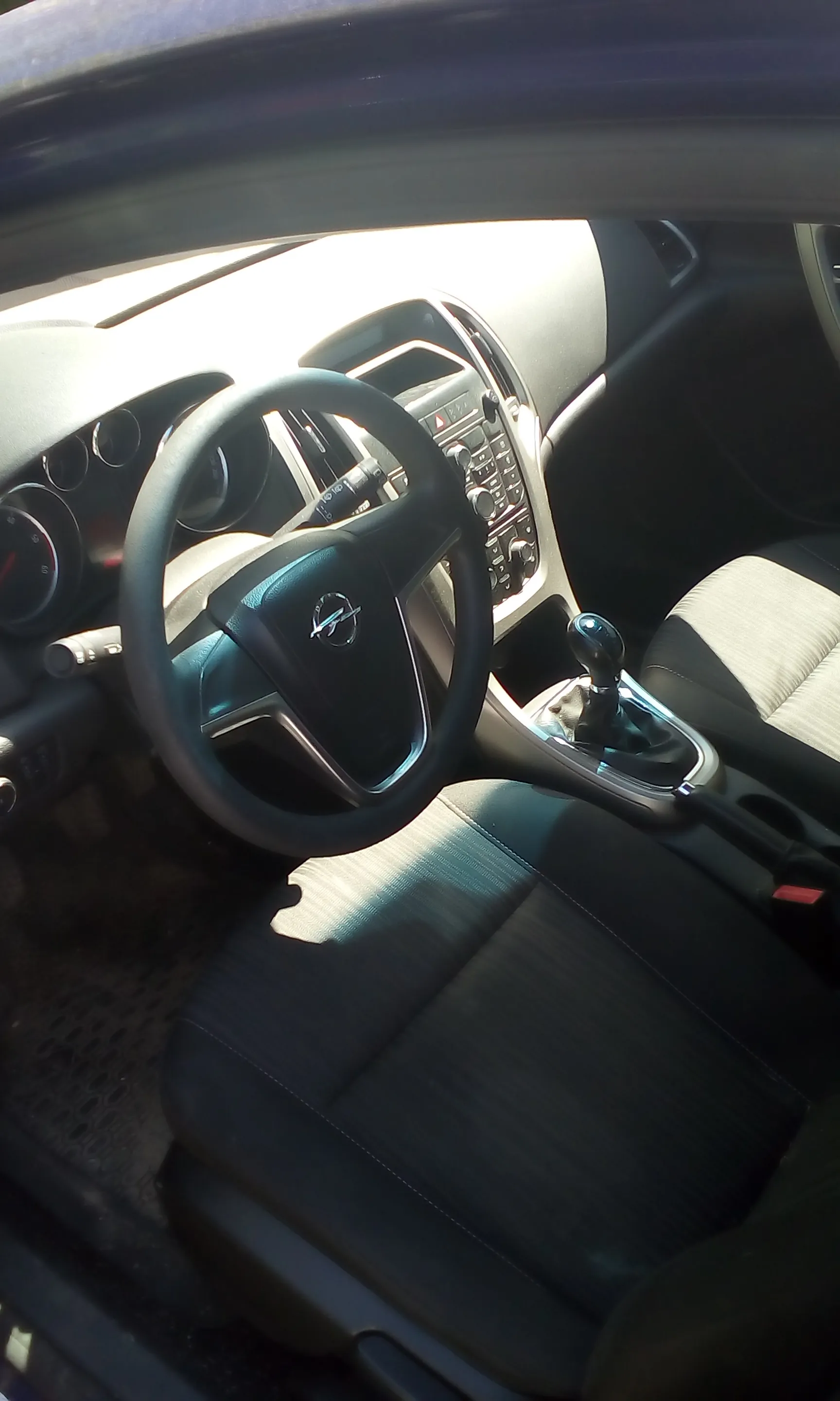 Wnętrze samochodu marki Opel widziane z perspektywy kierowcy. Widoczna jest czarna kierownica z logo producenta w centrum, deska rozdzielcza z systemem audio i klimatyzacją, wajcha zmiany biegów oraz ciemnoszare, materiałowe fotele. Zdjęcie wykonane z lekkiego kąta, z widocznym fragmentem ramy drzwi u góry.