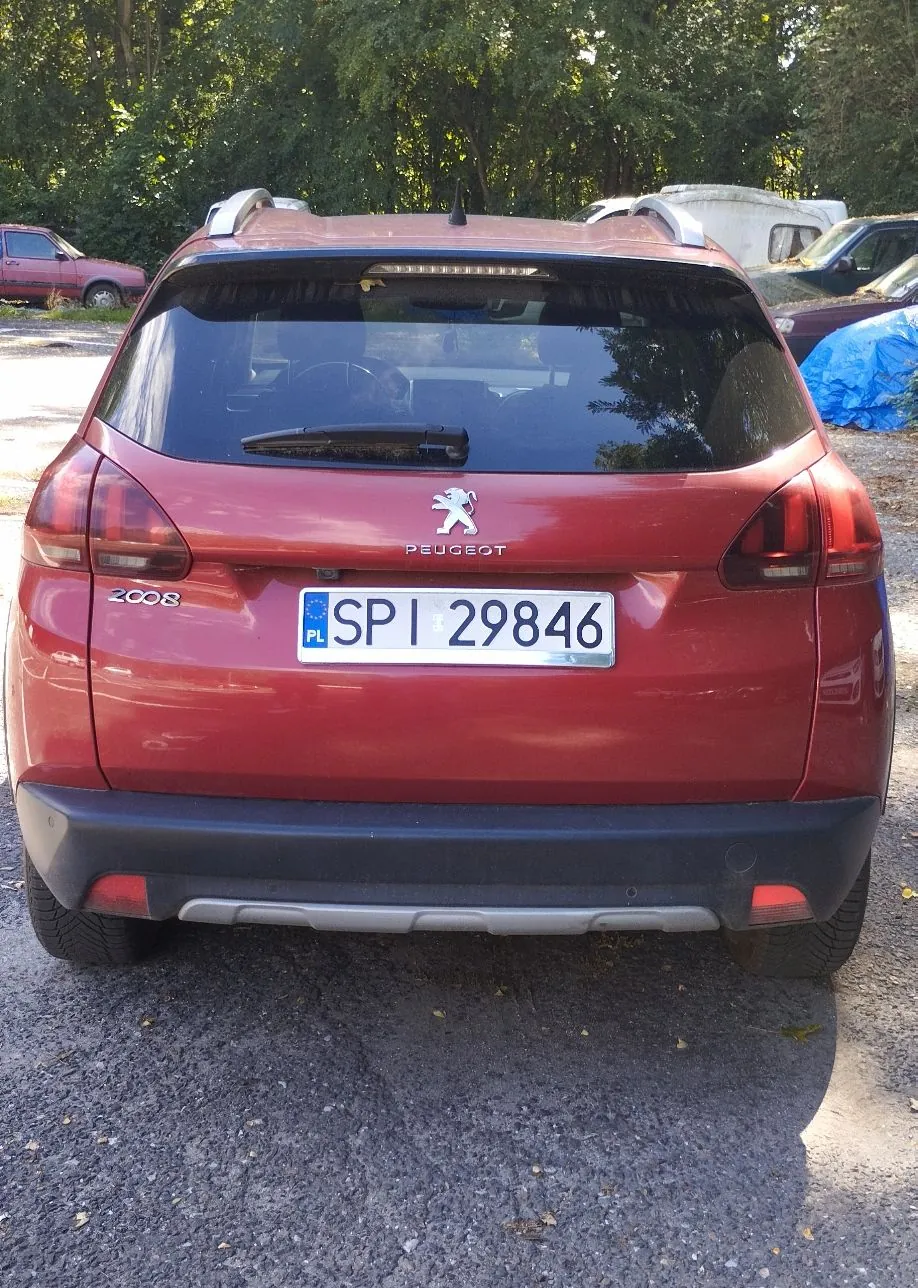 Tylny widok czerwonego samochodu marki Peugeot, model 2008. Pojazd posiada polską tablicę rejestracyjną o numerze SPI 29846. Na klapie bagażnika widoczne jest logo lwa oraz napis PEUGEOT. Samochód zaparkowany jest na żwirowym podłożu na tle zielonych drzew.