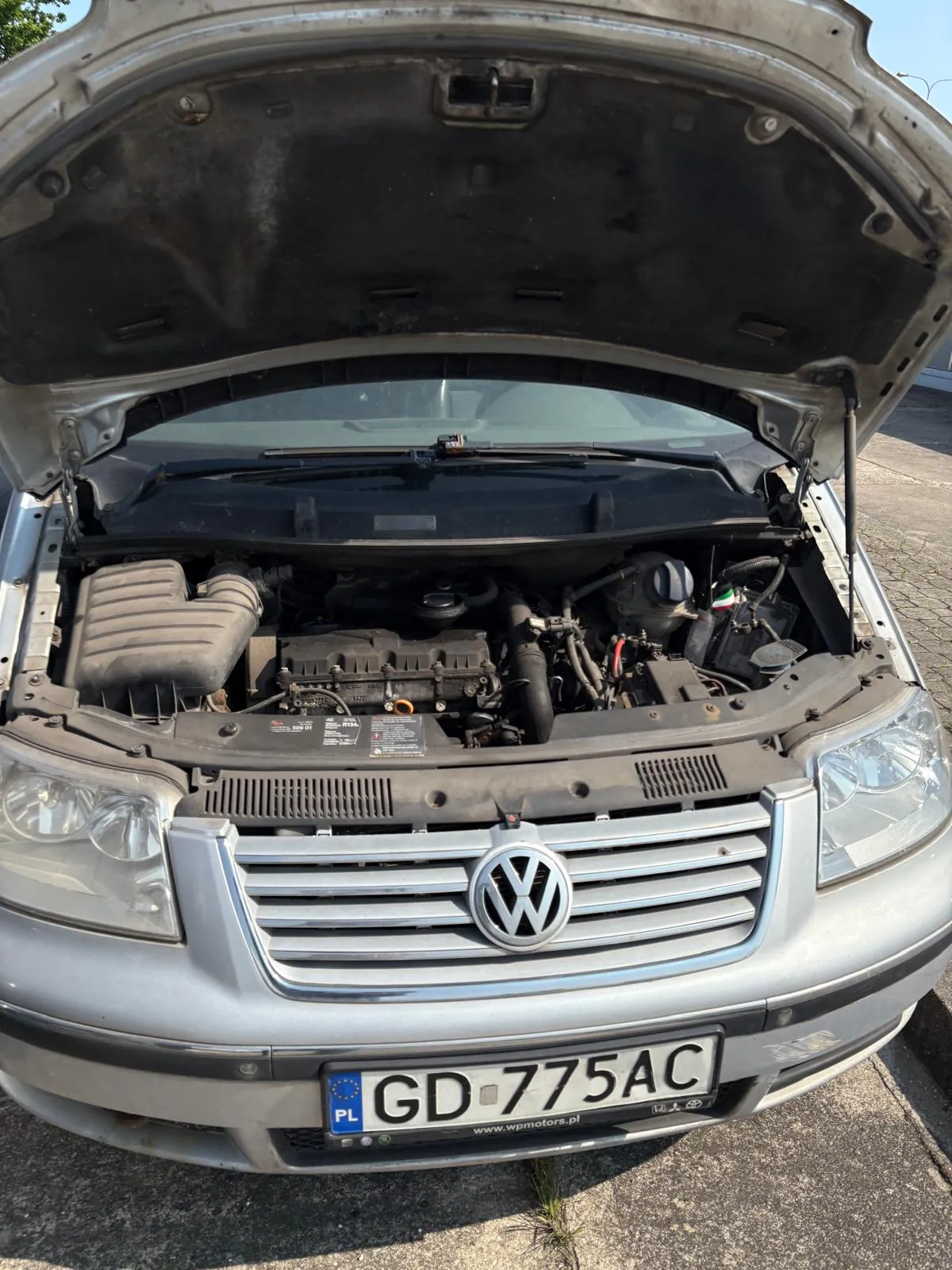 Przednia część srebrnego samochodu marki Volkswagen z otwartą maską. Widoczny silnik, elementy układu chłodzenia i filtr powietrza. Na masce umieszczony jest logo VW. Poniżej znajduje się tablica rejestracyjna z polskim kodem PL i numerem GD-775AC oraz adres strony www.wpmotors.pl. Tło stanowi asfaltowa powierzchnia i fragment zieleni.