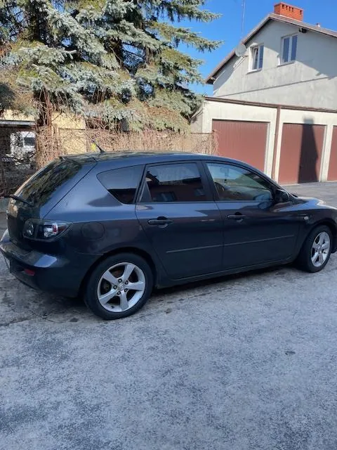 Ciemnoszary samochód typu hatchback zaparkowany na betonowej powierzchni. W tle dom z czerwonymi bramami garażowymi, kominkiem oraz drzewem o niebieskawo-zielonych igłach. Jasne niebo bez chmur.