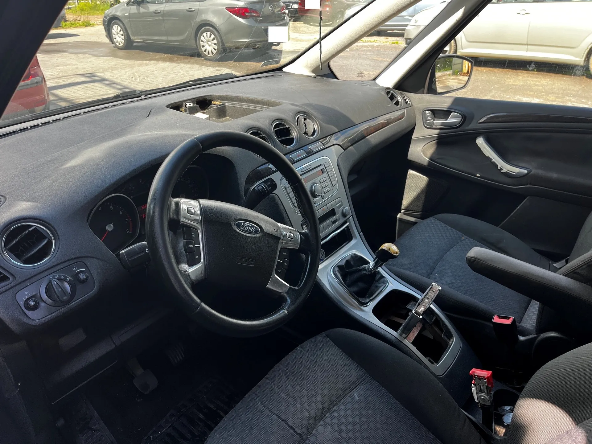 Wnętrze samochodu marki Ford, widok z perspektywy kierowcy. Widoczna czarna kierownica z logo Ford, deska rozdzielcza z systemem audio i nawiewami, czarne materiałowe fotele oraz lewarek zmiany biegów. Przez przednią szybę widoczne są inne zaparkowane samochody na placu.