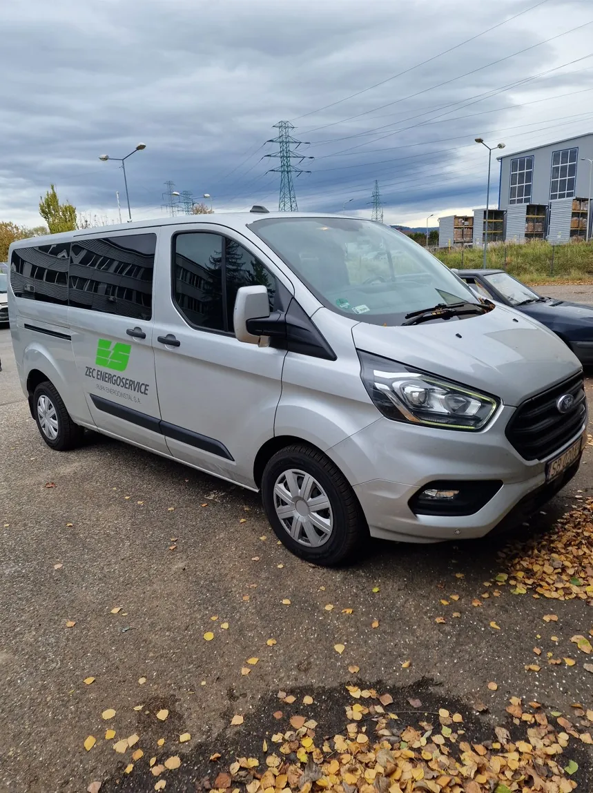 Srebrny samochód dostawczy marki Ford zaparkowany na asfaltowym placu z opadłymi żółtymi liśćmi. Na boku pojazdu znajduje się zielone logo oraz napis: 'ZEC ENERGOSERVICE' i mniejszy tekst poniżej: 'SPÓŁKA AKCYJNA ENERGOWALOR S.A.'. W tle widoczne są słupy wysokiego napięcia, budynek biurowy oraz pochmurne niebo.