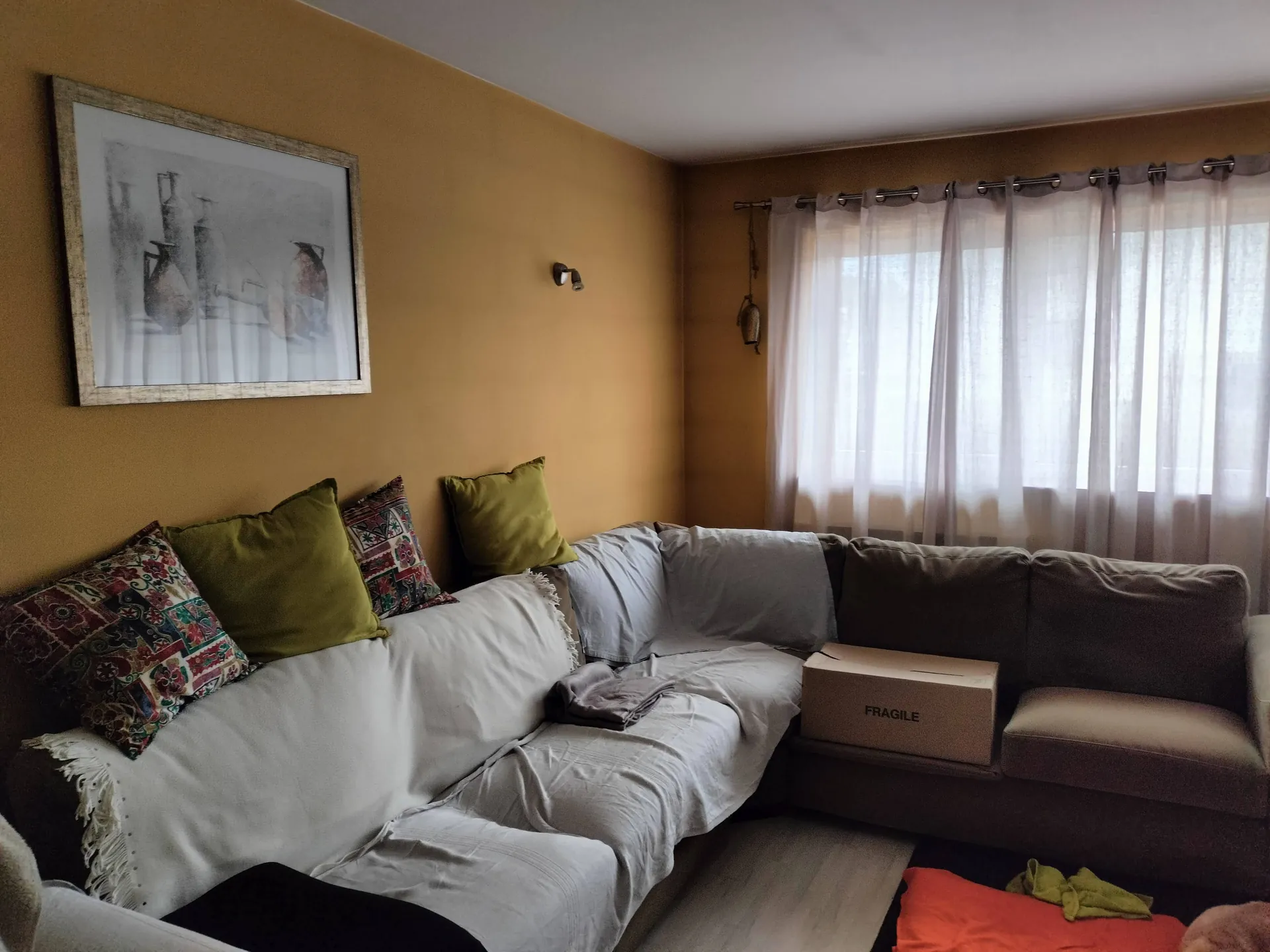 Wnętrze pokoju z brązową ścianą, na której wisi oprawiony w drewnianą ramę obraz przedstawiający martwą naturę z naczyniami. Poniżej znajduje się duża, narożna sofa przykryta białym prześcieradłem, z czterema poduszkami: dwiema zielonymi i dwiema w kolorowe, etniczne wzory. Na sofie leży szary koc oraz kartonowy karton z napisem „FRAGILE”. W tle widoczne jest okno z białymi, półprzezroczystymi zasłonami.