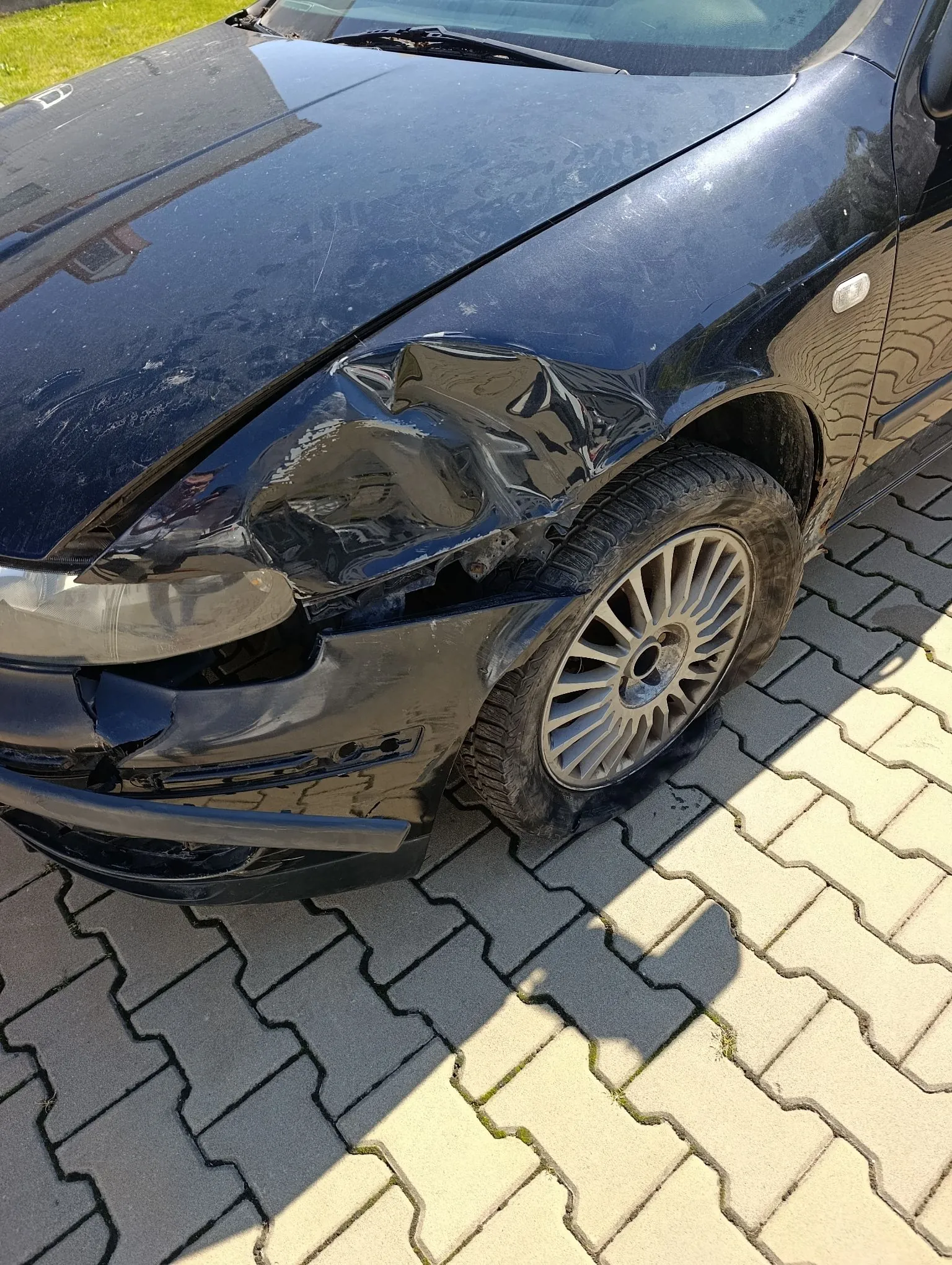 Zbliżenie na przód czarnego samochodu z poważnymi uszkodzeniami karoserii po lewej stronie. Widoczne jest wgniecenie maski, rozbite reflektory oraz całkowicie przebite przednie lewe koło, które opiera się o kostkę brukową. Samochód stoi na jasnoszarej kostce brukowej.