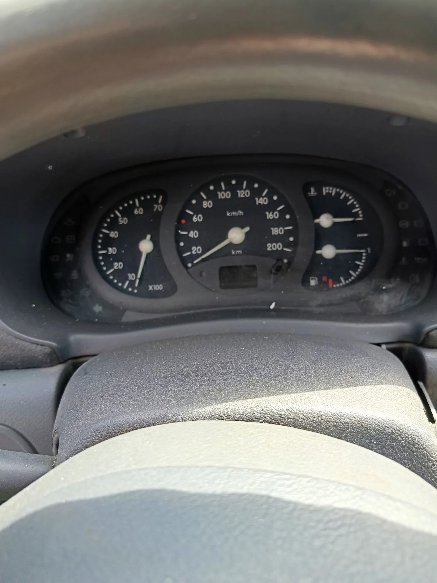 Zdjęcie deski rozdzielczej samochodu z widocznymi analogowymi wskaźnikami. W centrum znajduje się prędkościomierz z podziałką do 200 km/h, po lewej obrotomierz z oznaczeniem x100, a po prawej wskaźnik poziomu paliwa i temperatury. Poniżej prędkościomierza znajduje się mały, ciemny wyświetlacz cyfrowy. Widoczna jest również część kierownicy w dolnej i górnej części kadru.