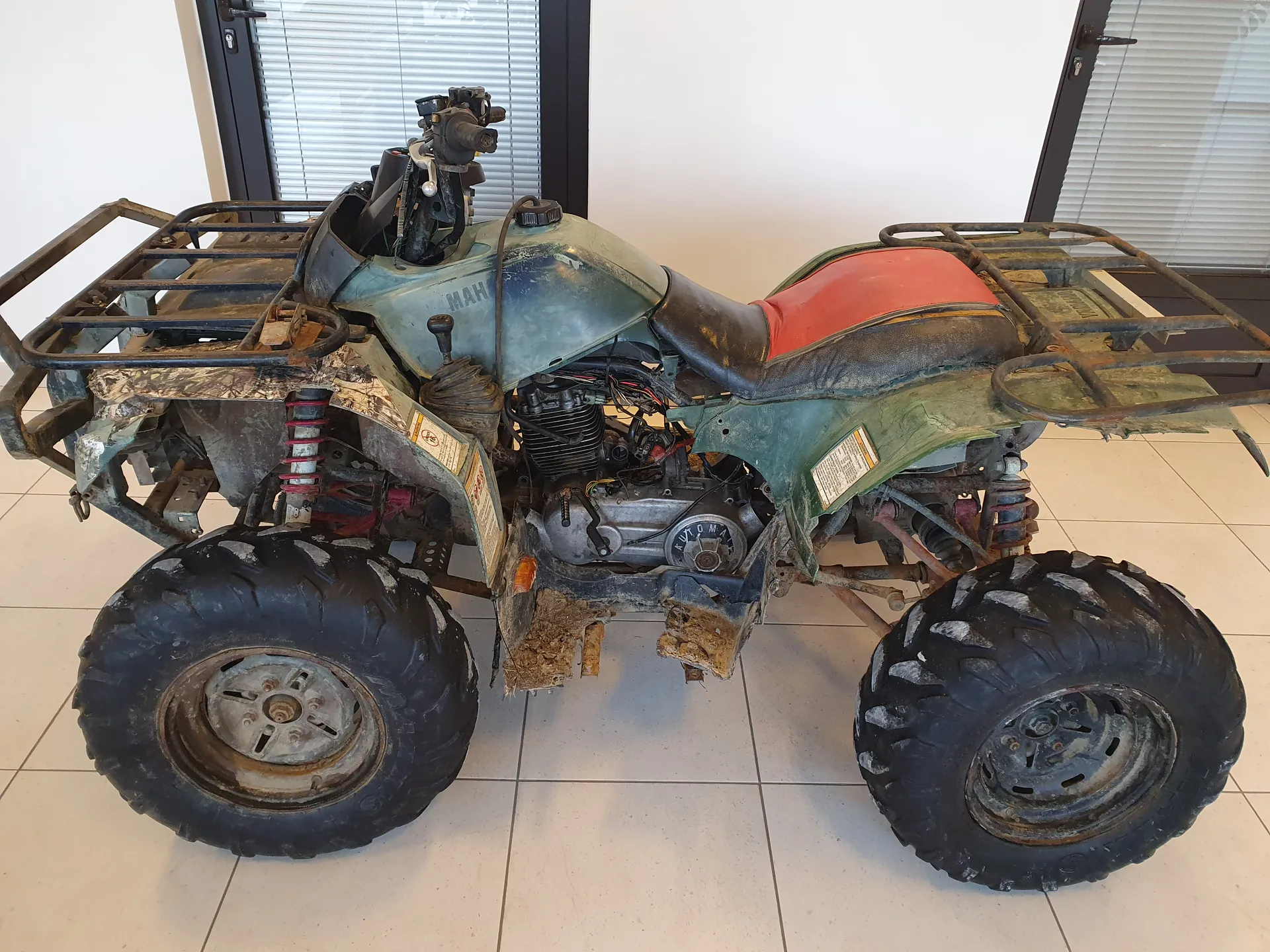 Zniszczony czterokołowy quad w kolorze zielonym z czerwonym siedzeniem, pokryty błotem i rdzą. Widoczne duże opony terenowe, metalowe rusztowania na przodzie i tył, odsłonięte elementy silnika oraz napisy: 'MAH' na zbiorniku paliwa i 'AUTOMAT' na silniku. Tło: wnętrze pomieszczenia z białą podłogą z płytek, białymi ścianami i oknami z żaluzjami.