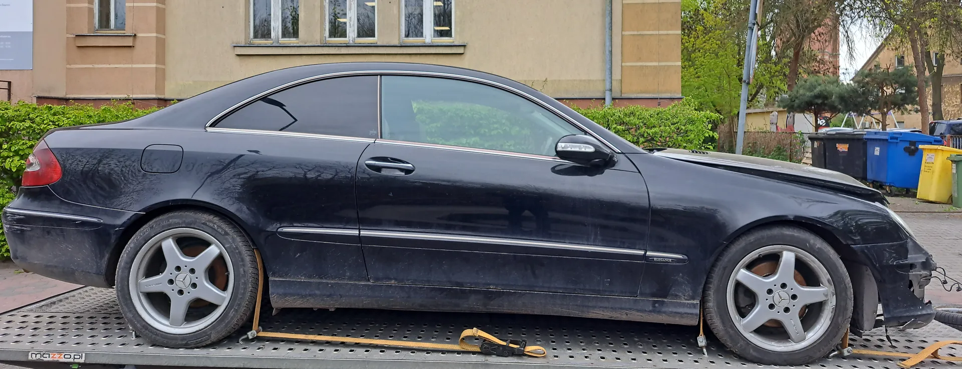 Czarny samochód osobowy marki Mercedes-Benz klasy CLK w wersji coupe, uszkodzony na przodzie (pęknięty zderzak, zdeformowana maska), umocowany żółtymi pasami na platformie holownika. Widoczne srebrne felgi z logo Mercedes-Benz, brudna karoseria. W tle budynki o beżowej elewacji z oknami, zielone krzewy, drzewa oraz kolorowe pojemniki na śmieci (czarny, niebieski, żółty, zielony). Na platformie holownika widoczny napis 'mazzo.pl'.