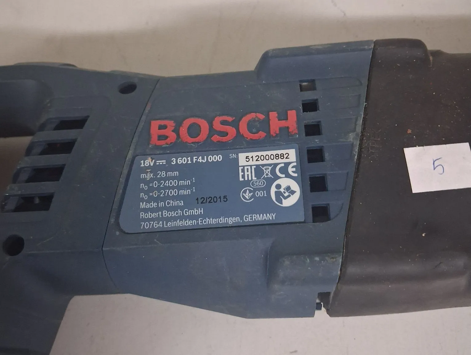 Zbliżenie na niebieską obudowę elektronarzędzia marki Bosch z naklejoną tabliczką znamionową. Na tabliczce znajduje się czerwony napis BOSCH oraz dane techniczne: 18V, numer modelu 3 601 F4J 000, numer seryjny SN: 512000882, maksymalna średnica 28 mm, prędkość obrotowa n0 = 0-2400 min-1 oraz n0 = 0-2700 min-1. Widnieje również napis Made in China, data 12/2015, nazwa firmy Robert Bosch GmbH oraz adres 70764 Leinfelden-Echterdingen, GERMANY. Na etykiecie znajdują się znaki certyfikacji EAC i CE oraz piktogram nakazujący używanie okularów ochronnych.
