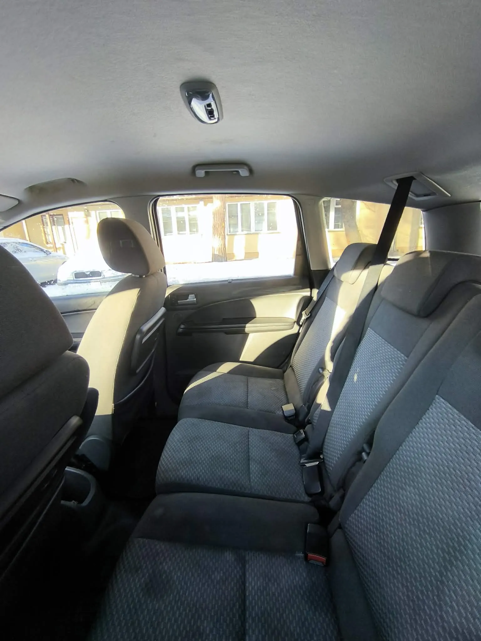 Wnętrze samochodu widziane z tyłu. Widoczne są czarne siedzenia z wzorzystą tapicerką, pasy bezpieczeństwa, sufit z lusterkiem wstecznym i uchwytem. Przez przednią szybę widać budynek z oknami, drzewo oraz zaparkowany samochód. Na zewnątrz na ziemi leży śnieg, a słońce rzuca cienie do wnętrza pojazdu.