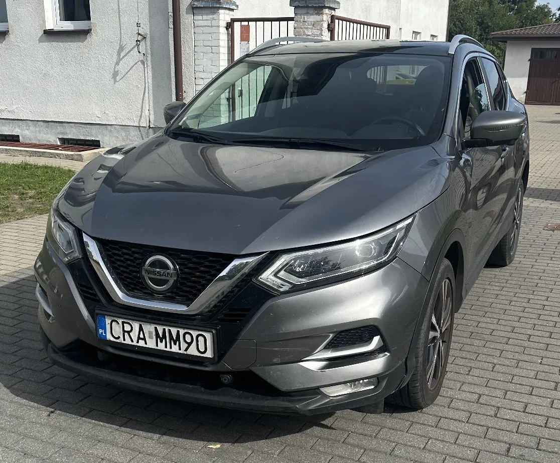 Srebrnoszary samochód marki Nissan zaparkowany na kostce brukowej przed białym budynkiem. Pojazd jest widoczny z przodu pod kątem, z wyraźnie widocznym logo Nissana na grillu oraz polską tablicą rejestracyjną o numerze CRA MM90.