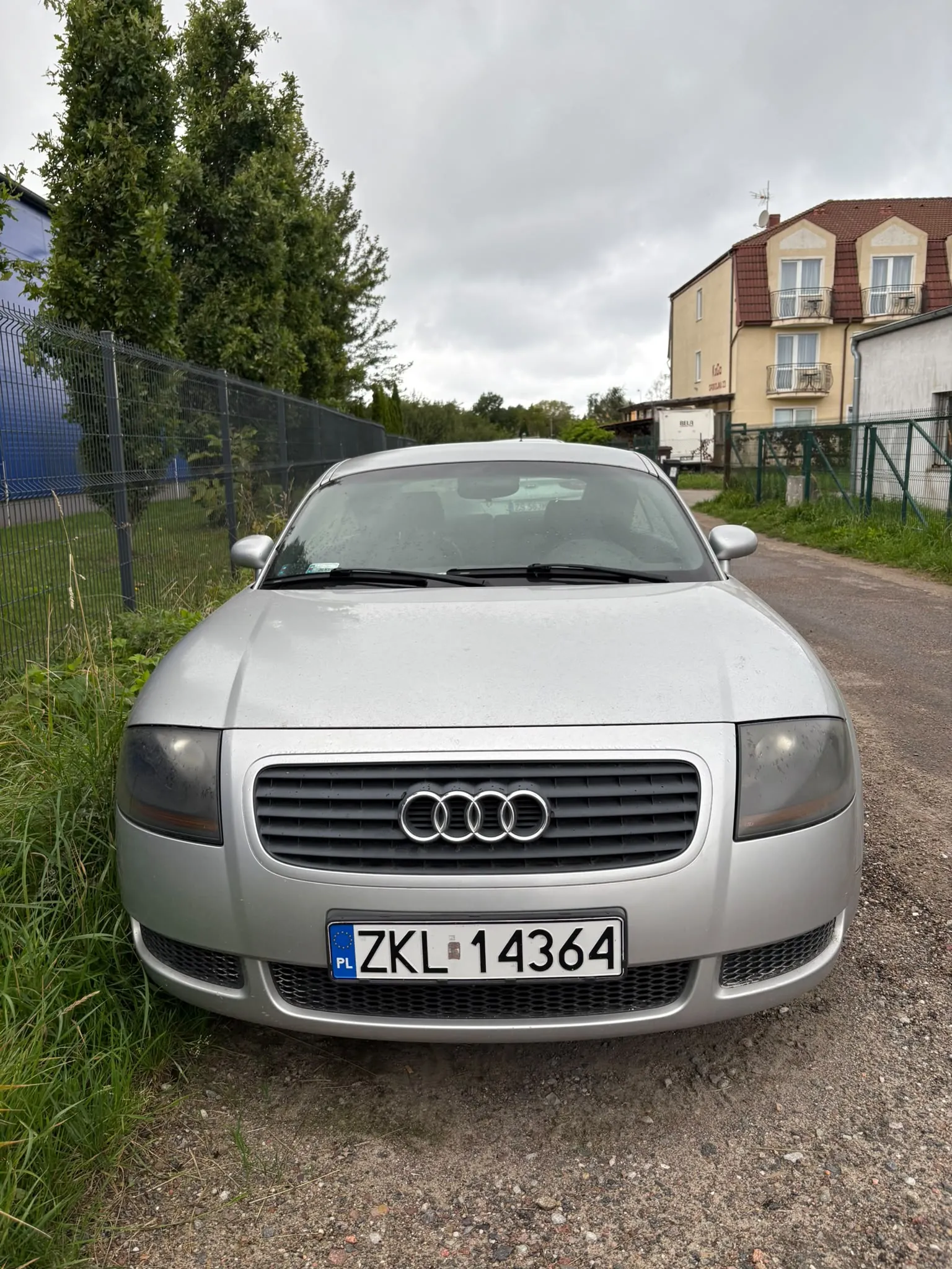 Srebrny samochód marki Audi zaparkowany na żwirowej drodze. Widoczny jest przód pojazdu z grillem, logo Audi oraz tablicą rejestracyjną o numerze ZKL 14364. W tle znajduje się zielony płot, drzewa oraz jasny, dwupiętrowy budynek mieszkalny pod zachmurzonym niebem.