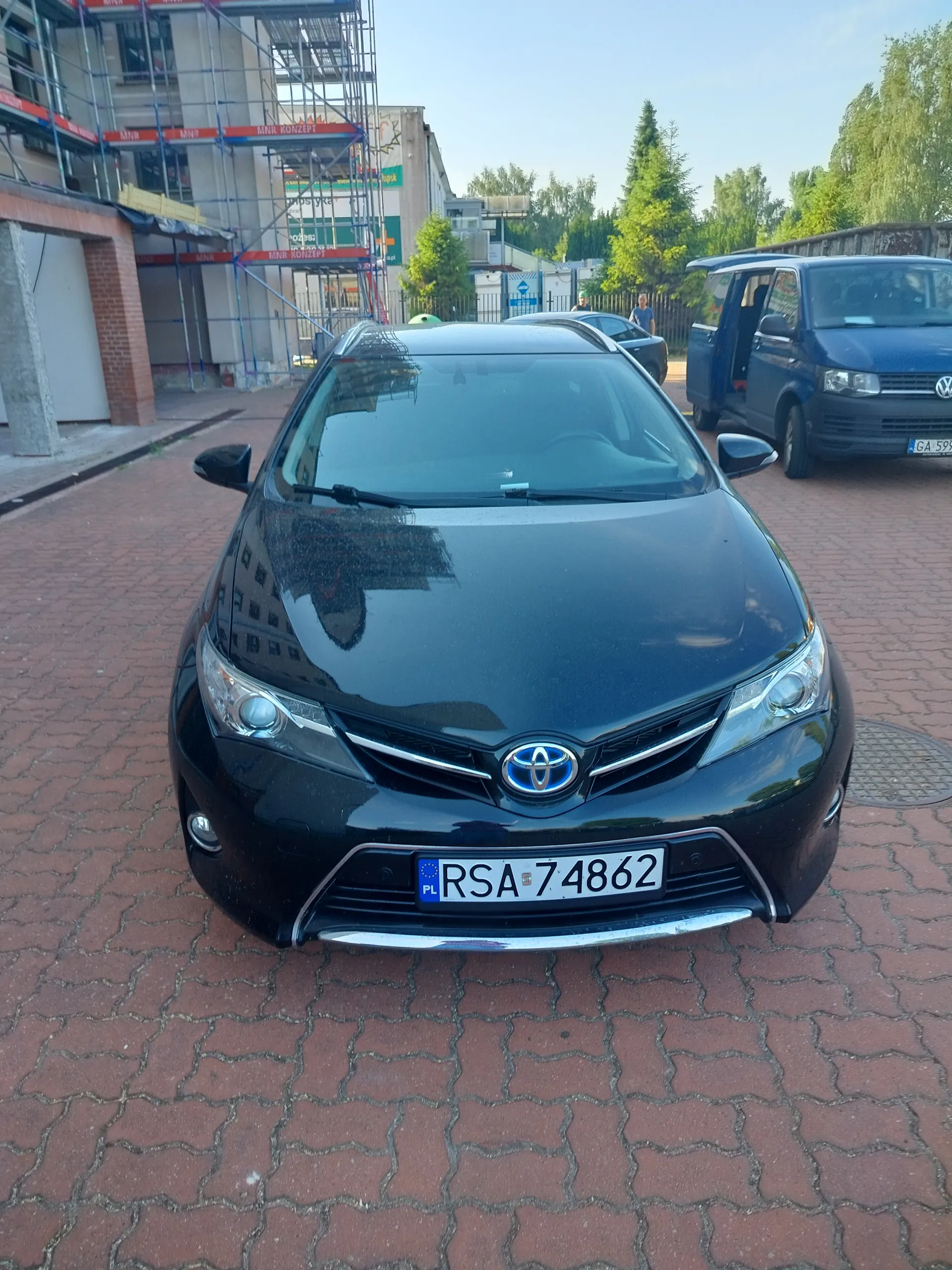 Czarny samochód marki Toyota z tablicą rejestracyjną PL RSA 74862 zaparkowany na czerwonej brukowanej nawierzchni. W tle budowla z rusztowaniem, na którym widoczne są czerwone napisy MIR KONCEPT, niebieski samochód dostawczy Volkswagen z otwartymi drzwiami oraz inne pojazdy. Po prawej stronie drzewa, a w oddali budynki i jasne niebo. Na tablicy rejestracyjnej samochodu widoczny jest niebieski pas z literami PL oraz białe tło z czarnymi znakami RSA 74862.
