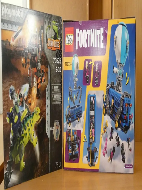 Dwa pudełka z zabawkami ustawione na drewnianym blacie. Po lewej stronie pudełko Playmobil z serii Dino Rise (numer 70626, wiek 5-10 lat, 73 elementy), przedstawiające zielonego dinozaura z konstrukcją i postaciami. Po prawej stronie pudełko LEGO Fortnite (numer 77073) z niebieskim balonem, pojazdem i minifigurekami. Na obu pudełkach widoczne są logotypy, numery zestawów oraz ilustracje produktów.