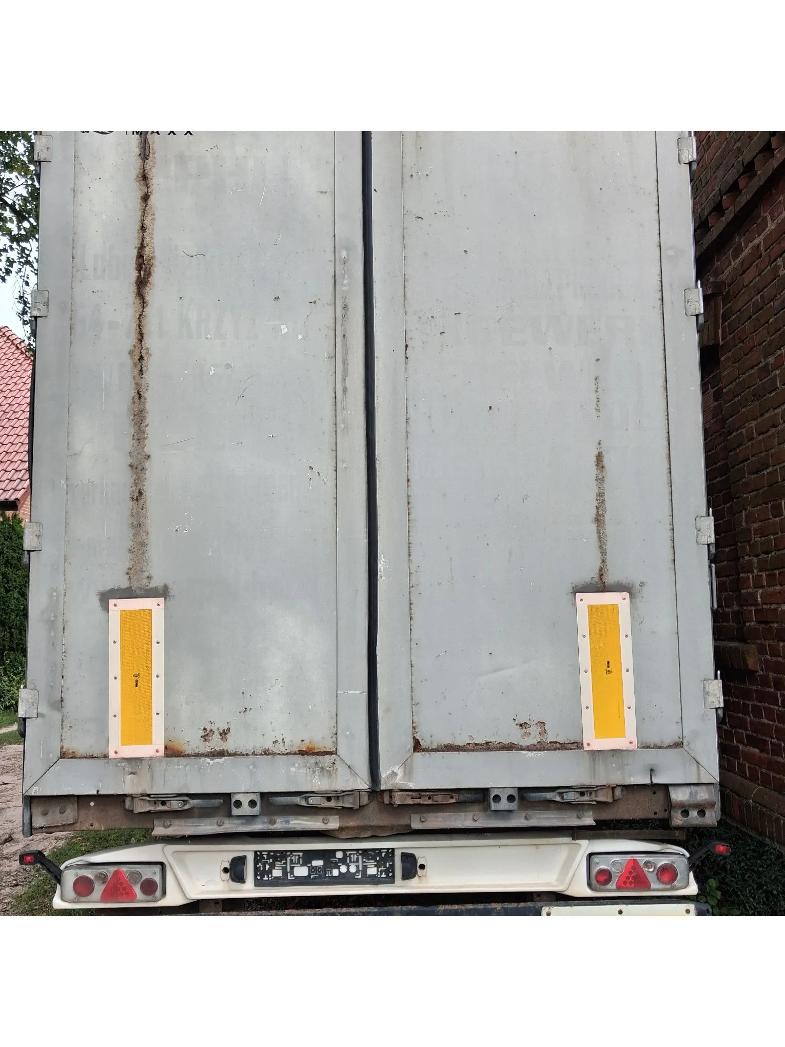 Tylny widok szarych, metalowych drzwi naczepy ciężarówki. Drzwi są lekko zardzewiałe, szczególnie wzdłuż krawędzi i łączeń. Na obu skrzydłach drzwi zamontowane są pionowe, żółte odblaski. Poniżej drzwi znajduje się biały zderzak z czerwonymi lampami tylnymi oraz tablicą rejestracyjną, która jest nieczytelna.