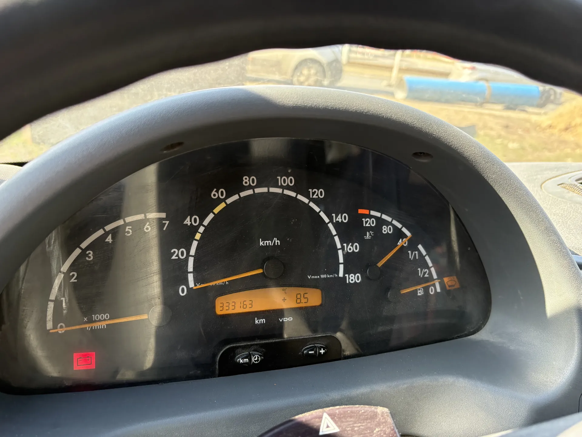 Zbliżenie na deski rozdzielcze samochodu. Widoczne są: wskaźnik prędkości (0-180 km/h), tachometr (0-7 x1000 1/min), wskaźnik paliwa (poziom około 1/2), termometr (0-120°C) oraz wyświetlacz cyfrowy z przebiegiem 333163 km i wartością 8.5. Na wyświetlaczu widoczne są oznaczenia 'km/h', 'Vmax 180 km/h', 'km', 'vdo'. Czerwony symbol akumulatora świeci się na lewo od tachometru. W tle przez przednią szybę rozmyty widok innego pojazdu i niebieskiego obiektu na terenie budowy.