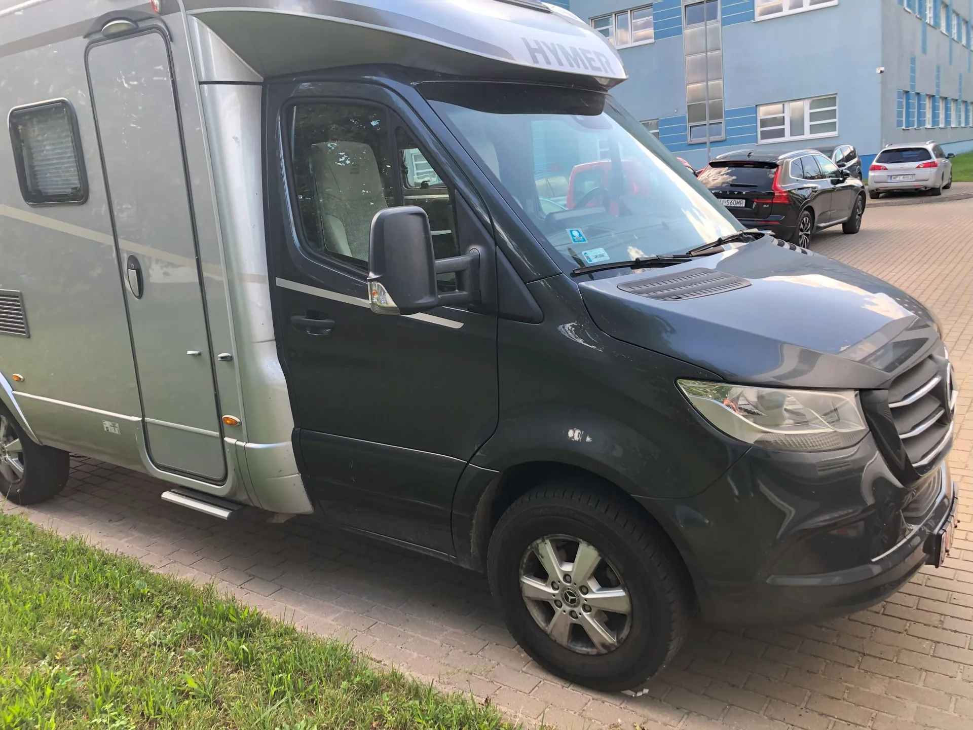 Srebrno-szary kamper marki Hymer oparty na podwoziu Mercedesa, zaparkowany na kostce brukowej obok trawnika. Pojazd ma ciemnoszarą kabinę kierowcy i srebrne nadwozie mieszkalne. W tle widoczny jest niebieski budynek biurowy oraz dwa zaparkowane samochody osobowe: czarny hatchback i biały SUV.