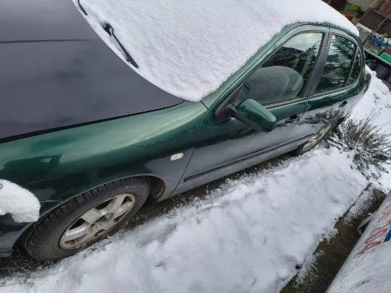 Zielony samochód osobowy zaparkowany na śniegu. Na dachu i przedniej szybie widoczna warstwa śniegu. Koła i dolna część nadwozia częściowo pokryte śniegiem. W tle fragment zasnieżonej ziemi i krzewów. Po prawej stronie obrazu widoczny fragment białego obiektu z czerwonymi literami, nieczytelnymi w całości.