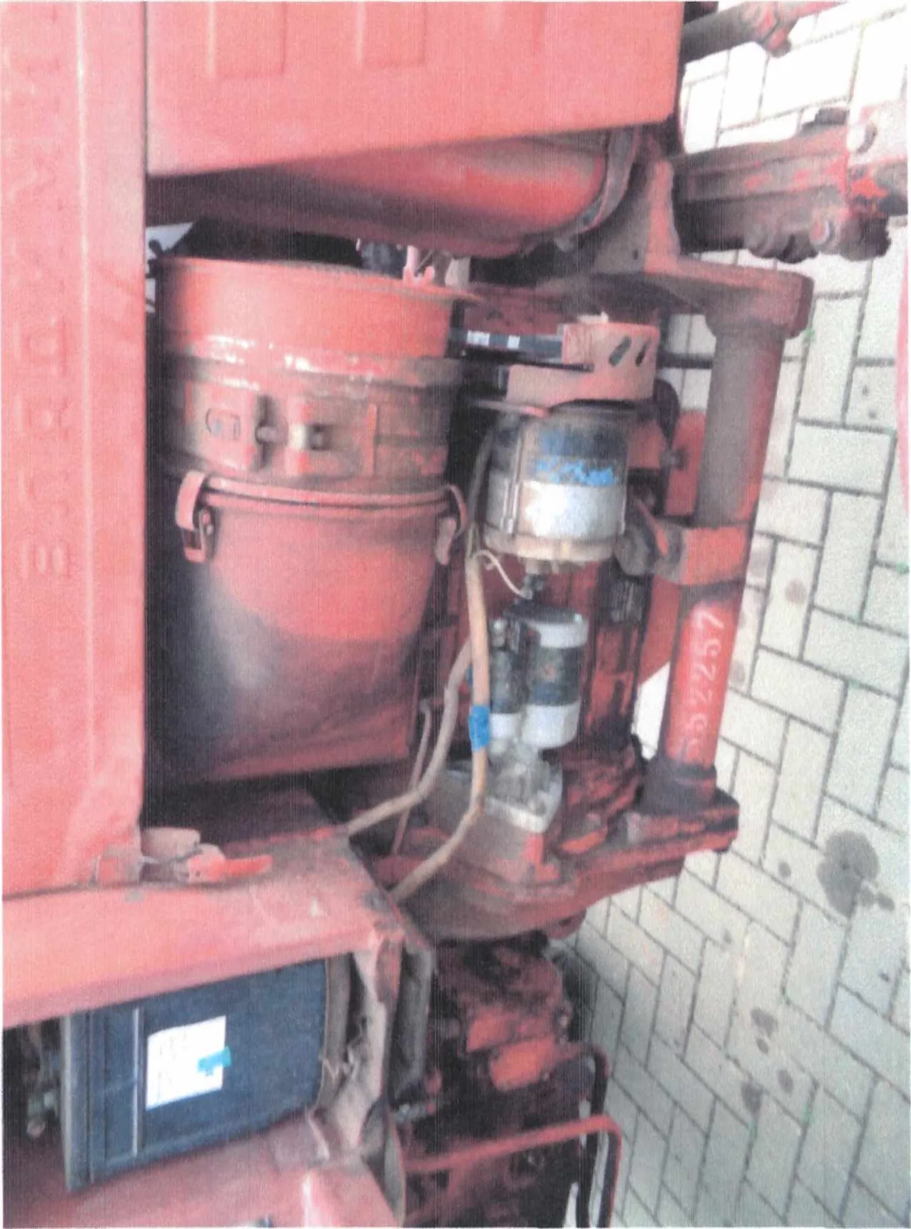 Zbliżenie na czerwony metalowy element maszyny lub pojazdu z widocznymi przewodami, cylindrycznym zbiornikiem i metalowymi częściami. Na dolnej części widoczny czerwony numer 552257. W tle niebieski pojemnik z białą etykietą i niebieskim znacznikiem oraz brukowana posadzka z kafelkami.