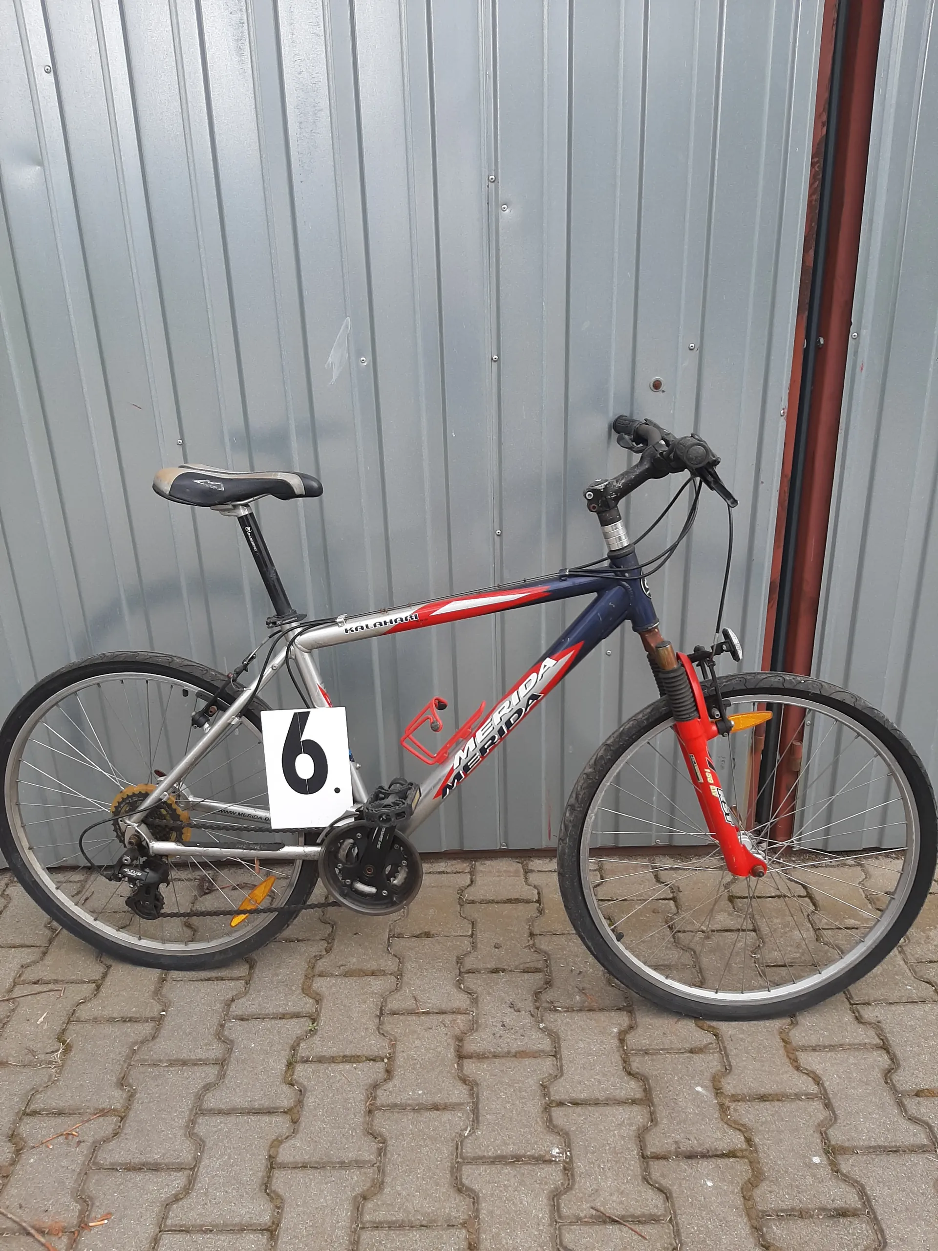 Rower o niebiesko-czerwonej ramie z widocznym napisem "MERIDA", czarnym siedziskiem, czerwonym uchwytem na bidon i białą tabliczką z czarną cyfrą "6". Rower oparty o metalową bramę garażową i brukowaną powierzchnię.