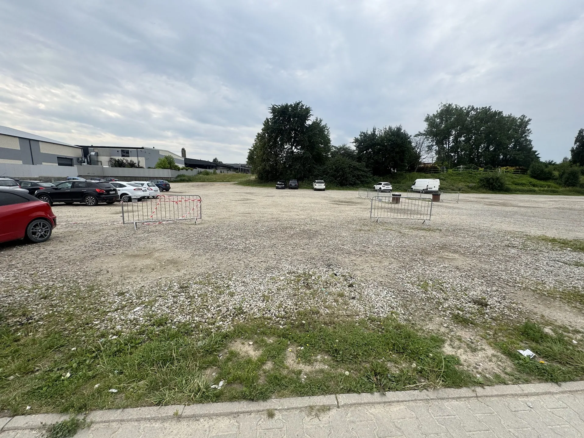 Szeroki widok na żwirowy parking pod pochmurnym niebem. Po lewej stronie zaparkowanych jest kilka samochodów, w tym czerwony samochód na pierwszym planie. W tle widoczne są niskie budynki przemysłowe oraz pas zieleni z drzewami. Na środku parkingu stoją dwie metalowe barierki odgradzające fragment terenu. W oddali widać kilka innych zaparkowanych pojazdów, w tym białego vana.