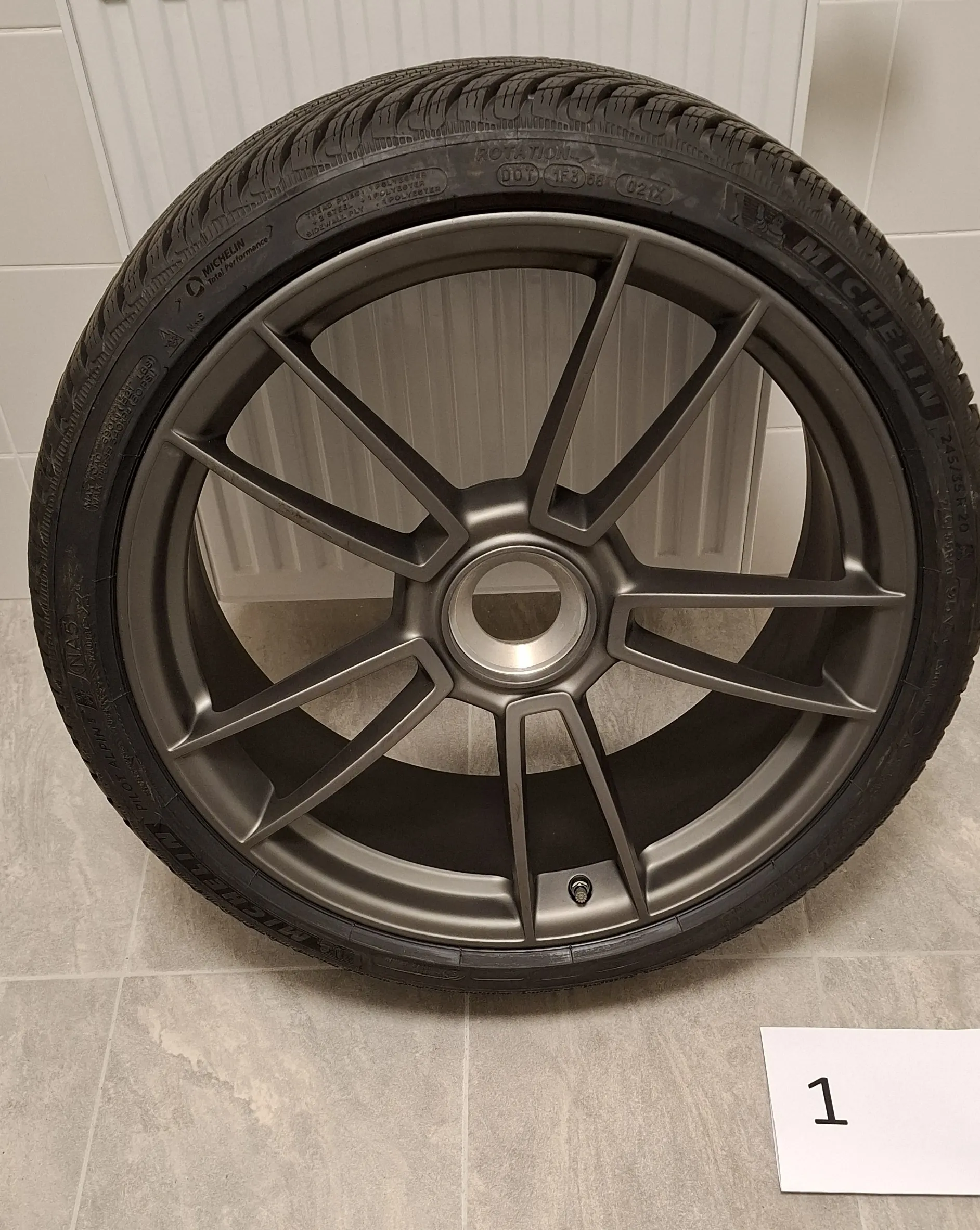 Zdjęcie koła samochodowego z oponą marki Michelin i ciemnoszarą, matową felgą aluminiową o pięciu podwójnych ramionach. Koło stoi pionowo na jasnej, płytkowej podłodze na tle białego grzejnika. W prawym dolnym rogu znajduje się biała kartka z czarną cyfrą 1.