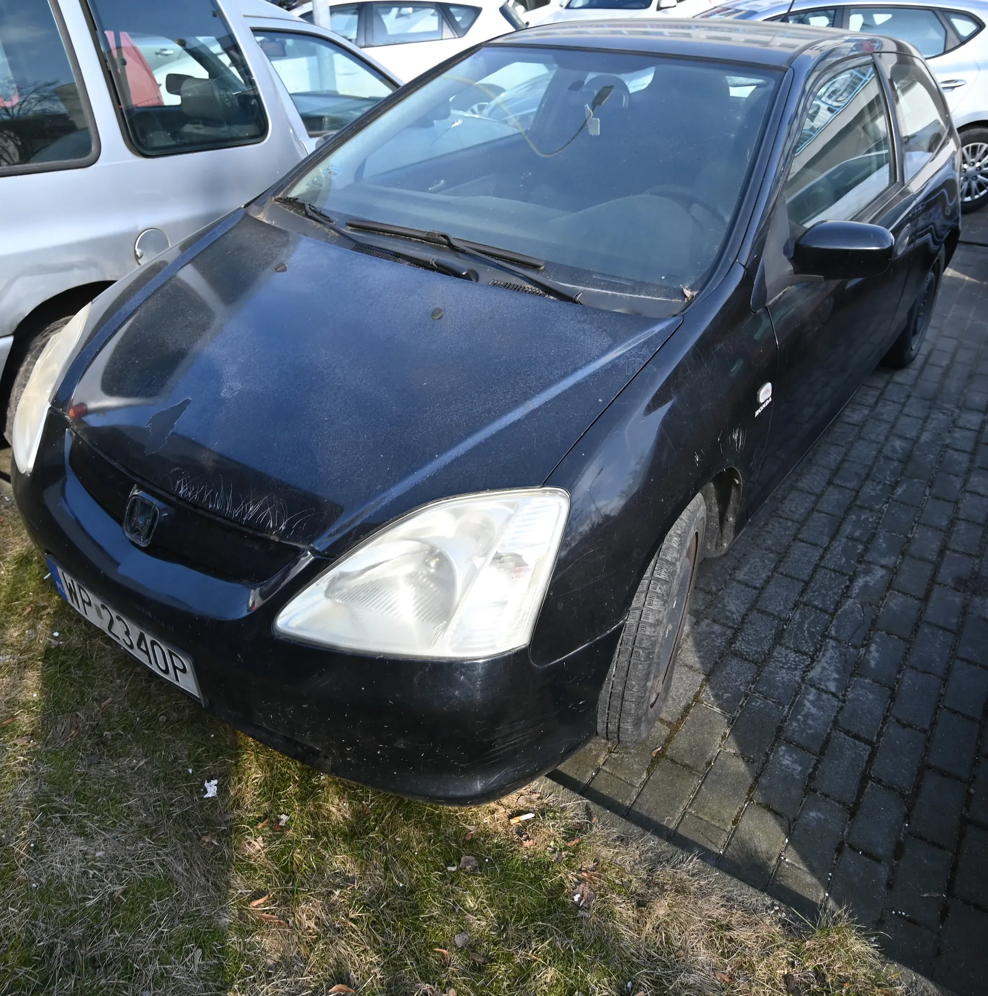 Czarny samochód marki Honda zaparkowany na brukowanej powierzchni. Widoczna tablica rejestracyjna WP 2340P. W tle inne pojazdy, w tym srebrny samochód. Podłoga składa się z bruku i trawnika z suchą trawą. Samochód ma lekko zabrudzoną karoserię i widoczne ślady uszkodzeń na masce.