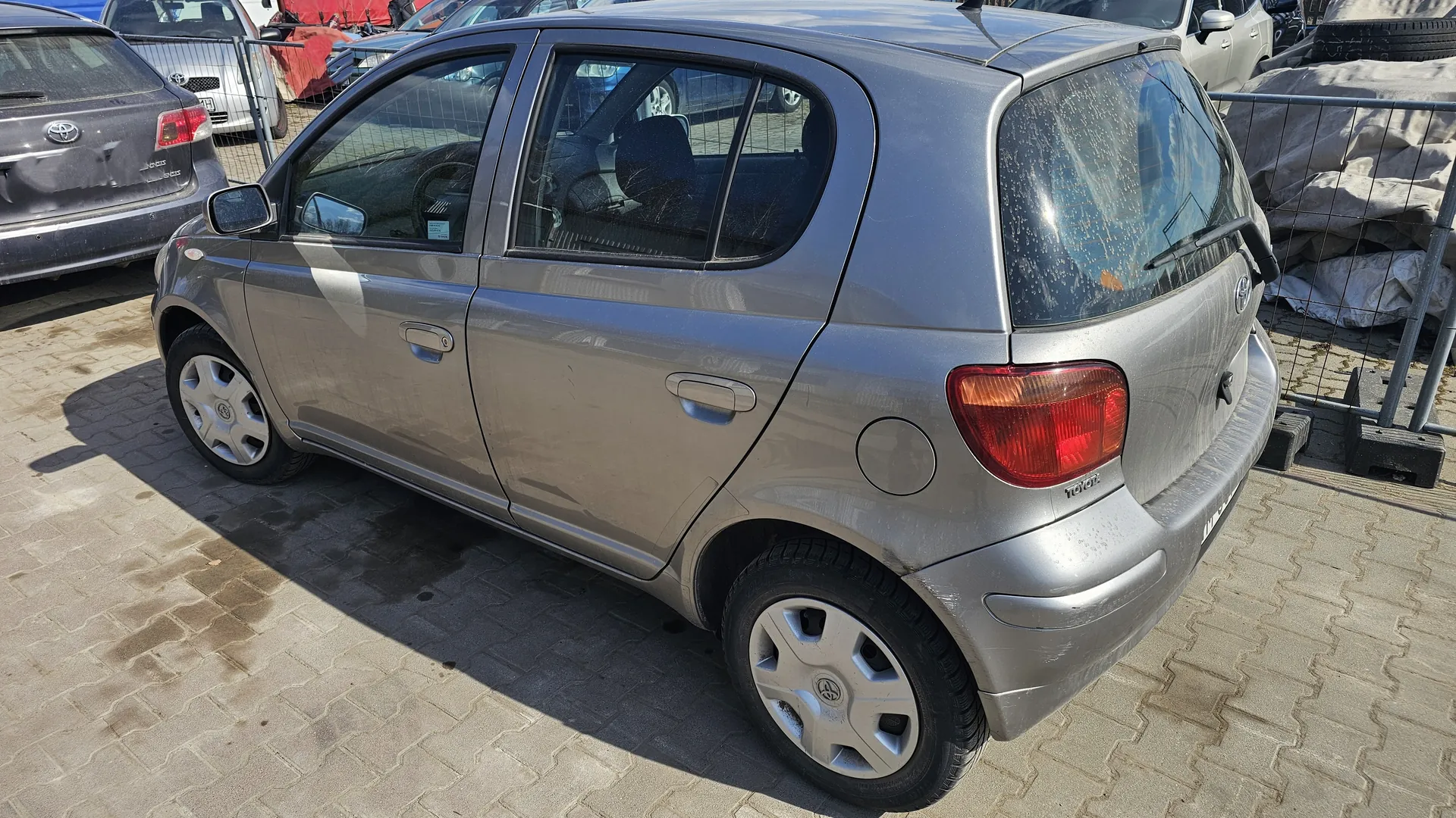 Srebrny samochód osobowy marki Toyota, widok z tyłu i boku. Pojazd zaparkowany na brukowanym placu. W tle widoczne inne samochody, metalowe ogrodzenie oraz zakryte materiałem przedmioty. Na tylnej szybie i nadwoziu widoczne ślady brudu. Logo Toyota umieszczone na tylnej części pojazdu. Koła z srebrnymi felgami z logo marki.
