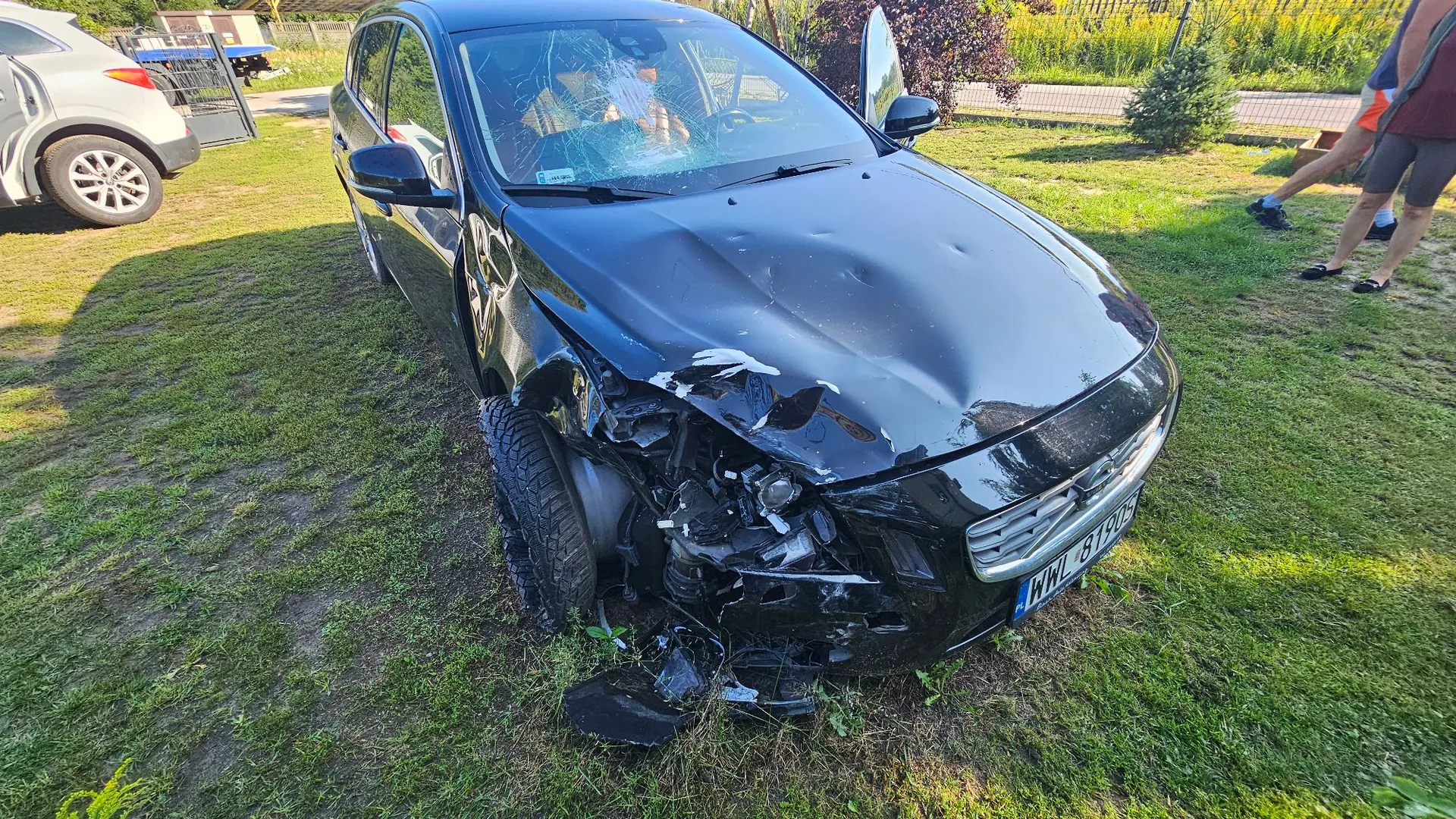 Czarny samochód osobowy z poważnymi uszkodzeniami przodu po kolizji. Maską jest wgnieciona, przedni zderzak i reflektory są zniszczone, a przednia szyba jest mocno rozbita. Pojazd stoi na trawiastym terenie. W tle widoczny jest fragment innego, białego samochodu oraz nogi osoby stojącej po prawej stronie. Tablica rejestracyjna samochodu to WWL 8190X.