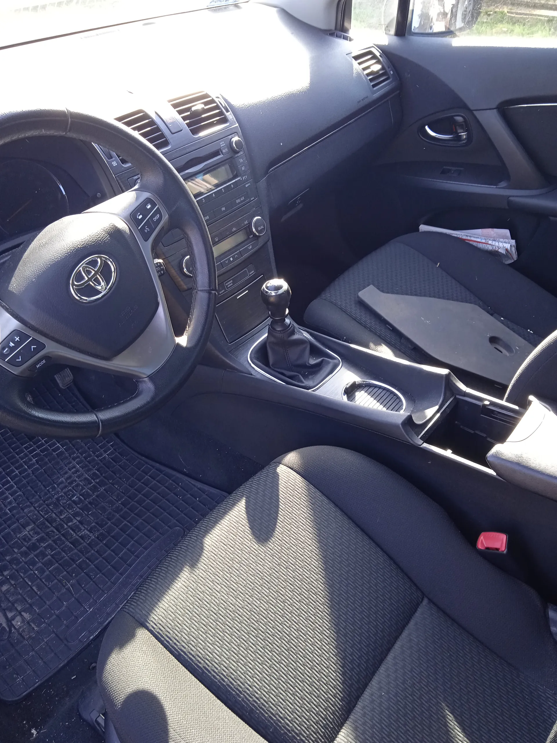 Wnętrze samochodu marki Toyota z widocznym kierownicą z logo, czarnym deski rozdzielczej z wentylacjami, radio i klimatyzacją. W centrum konsoli znajduje się dźwignia zmiany biegów, podłokietnik z uchwytem na kubek oraz czarne siedzenia z teksturą. Na podłodze pasażera leży gazeta, a na fotelu kierowcy widoczny jest czerwony zamek pasów bezpieczeństwa. Wnętrze oświetlone jest światłem słonecznym, rzucającym cienie na siedzenia.