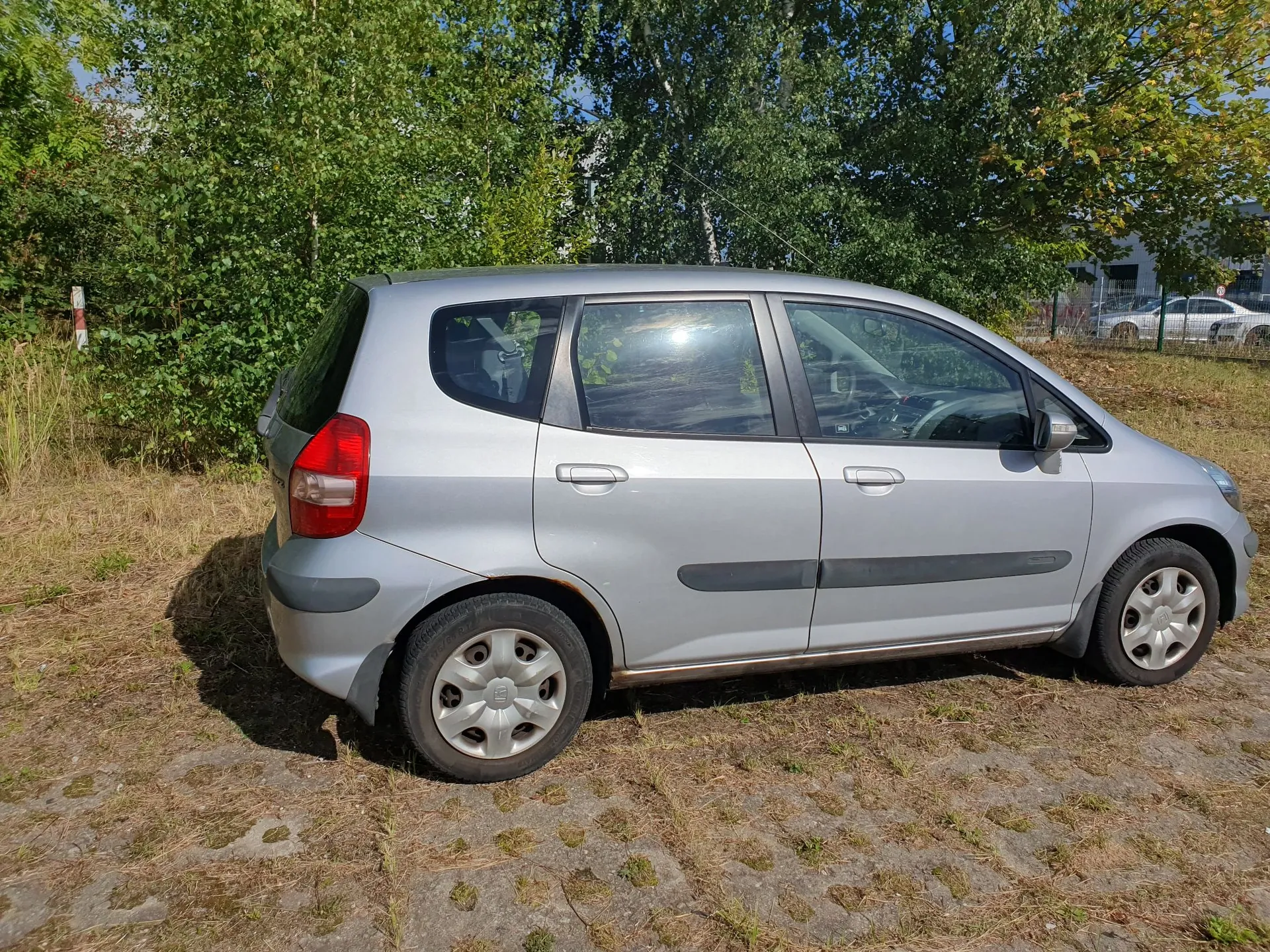 Srebrny samochód osobowy typu hatchback zaparkowany bokiem na utwardzonym placu z trawą. W tle widać gęste, zielone drzewa i krzewy oraz fragment białego samochodu w oddali.