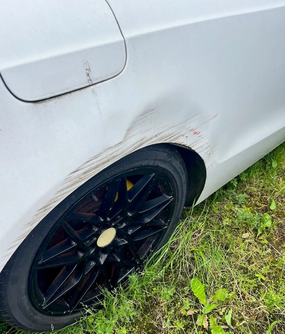 Zbliżenie na tylny prawy błotnik białego samochodu z widocznym wgnieceniem i zarysowaniami nad kołem. Felga samochodu jest czarna z żółtym logo w centrum. Auto stoi na trawie.